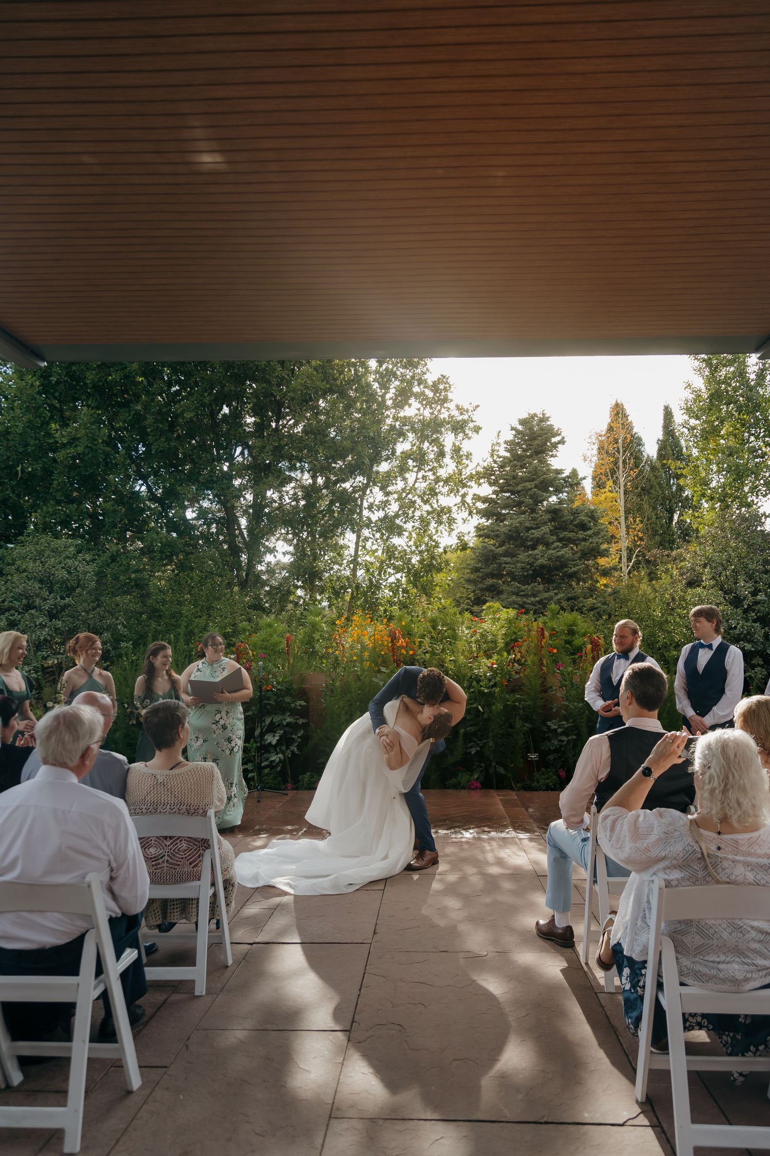 Bride and groom share first kiss at outdoor wedding ceremony with guests seated on white chairs.
