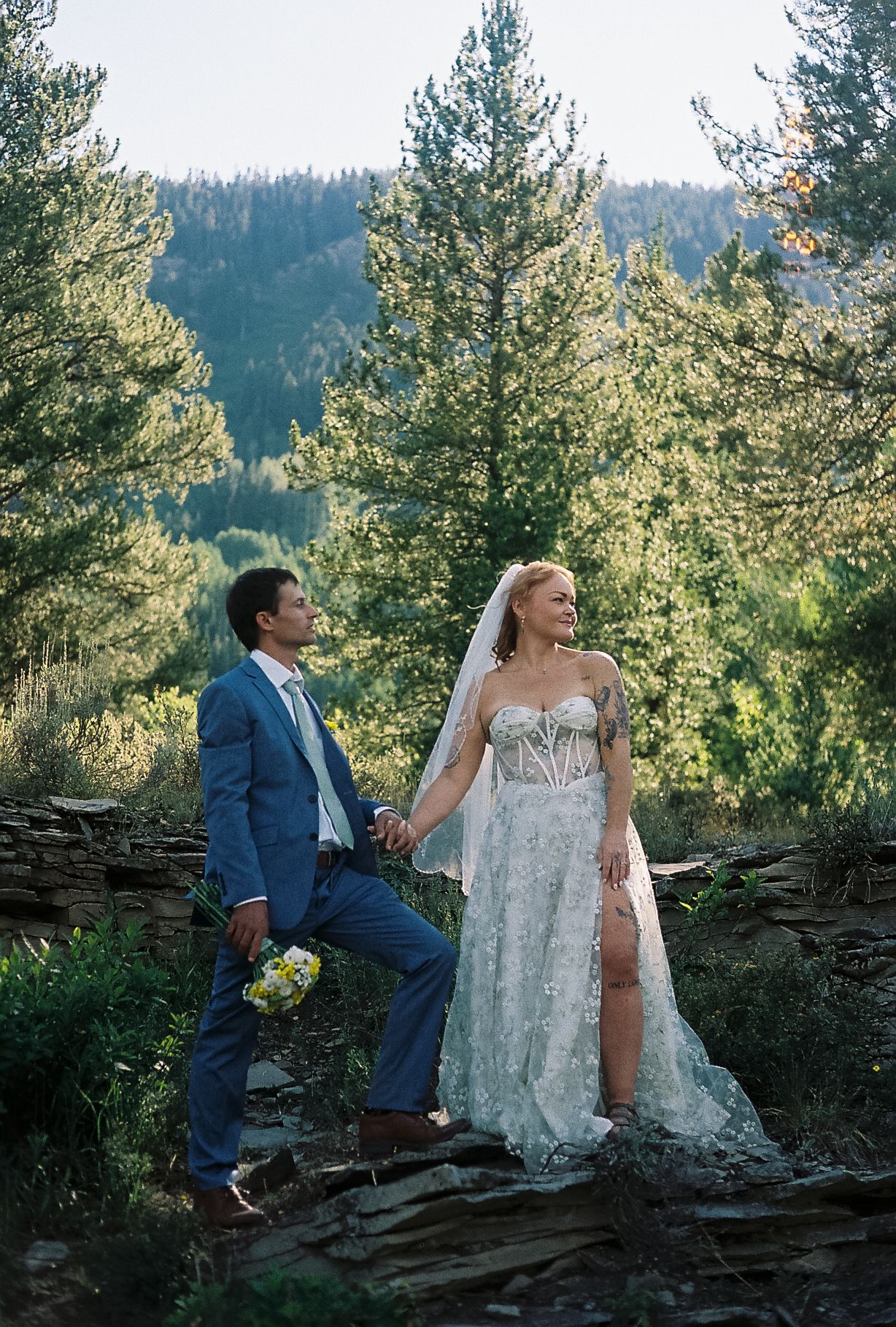 Bride and groom hold hands in a mountain forest setting, she in lace gown with veil, he in blue suit.