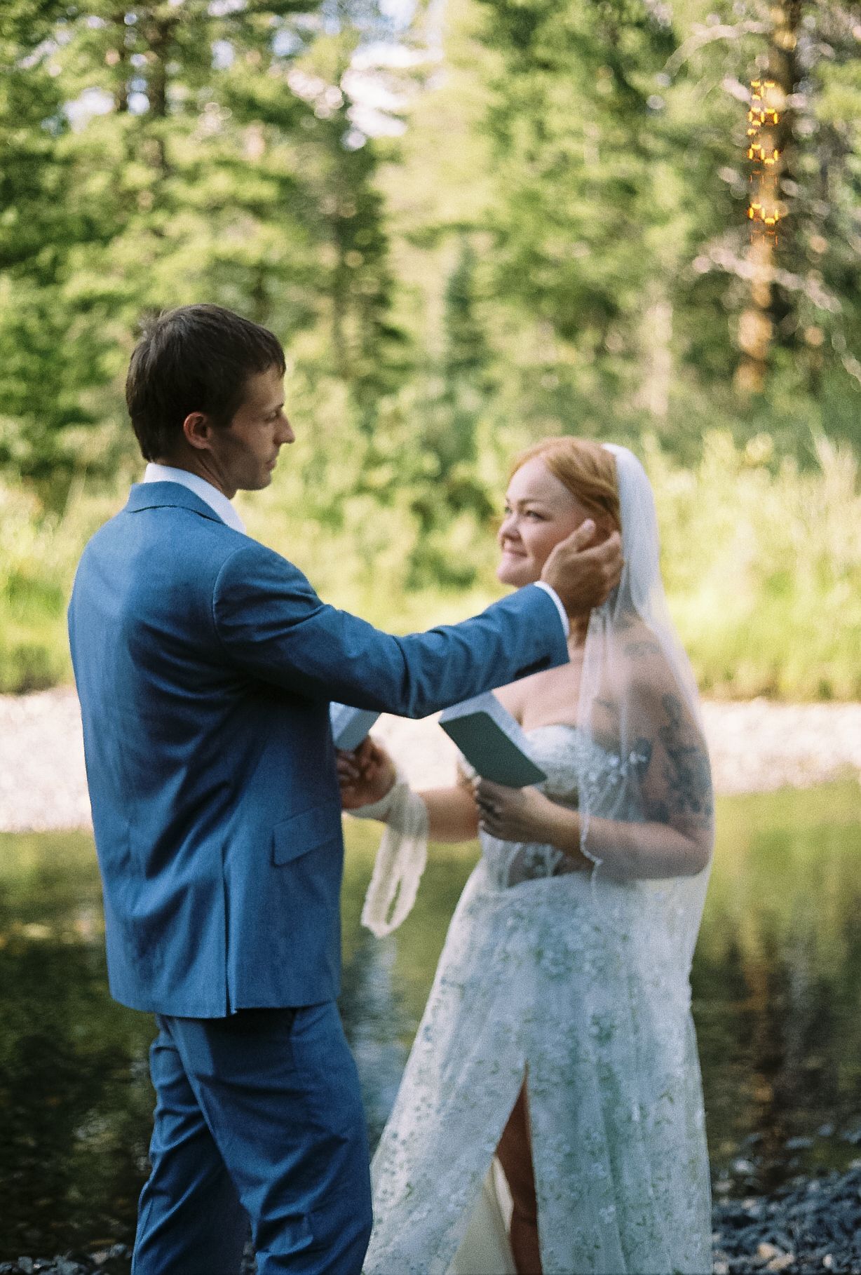 Groom tenderly brushes hair from brides face during outdoor vow exchange beside a forest stream.