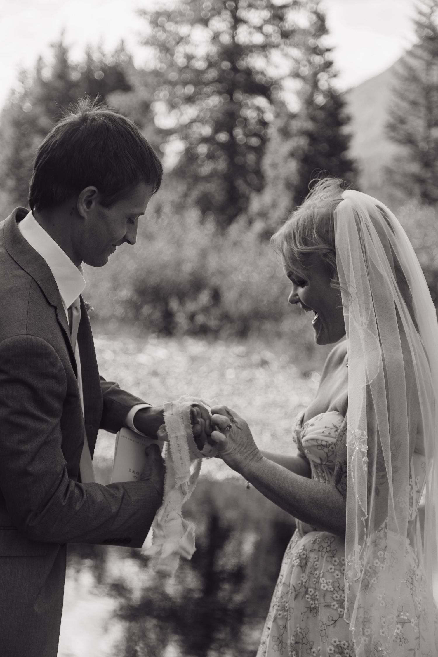 Black and white photo of groom placing wedding ring on bride's finger outdoors near trees.