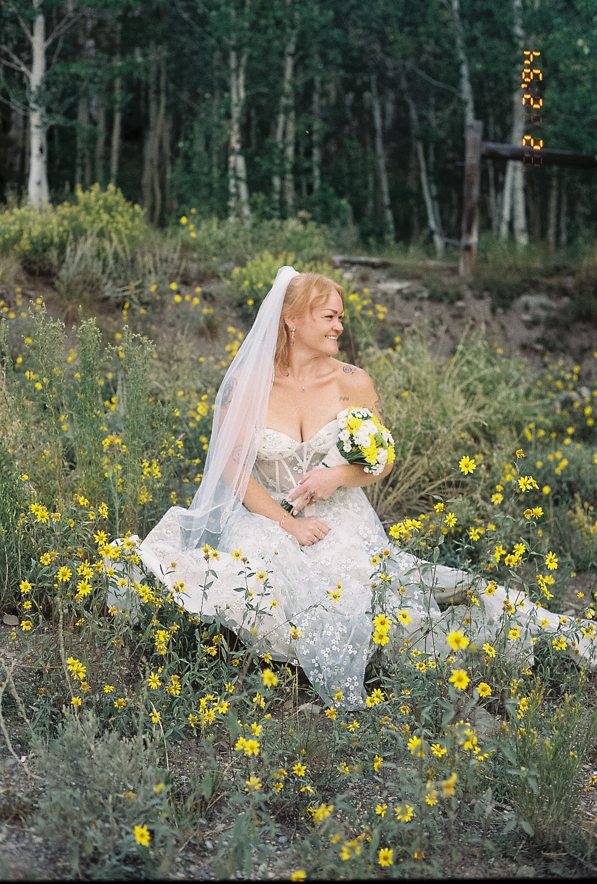 A smiling bride in a lace gown sits among yellow wildflowers holding a bouquet on film.
