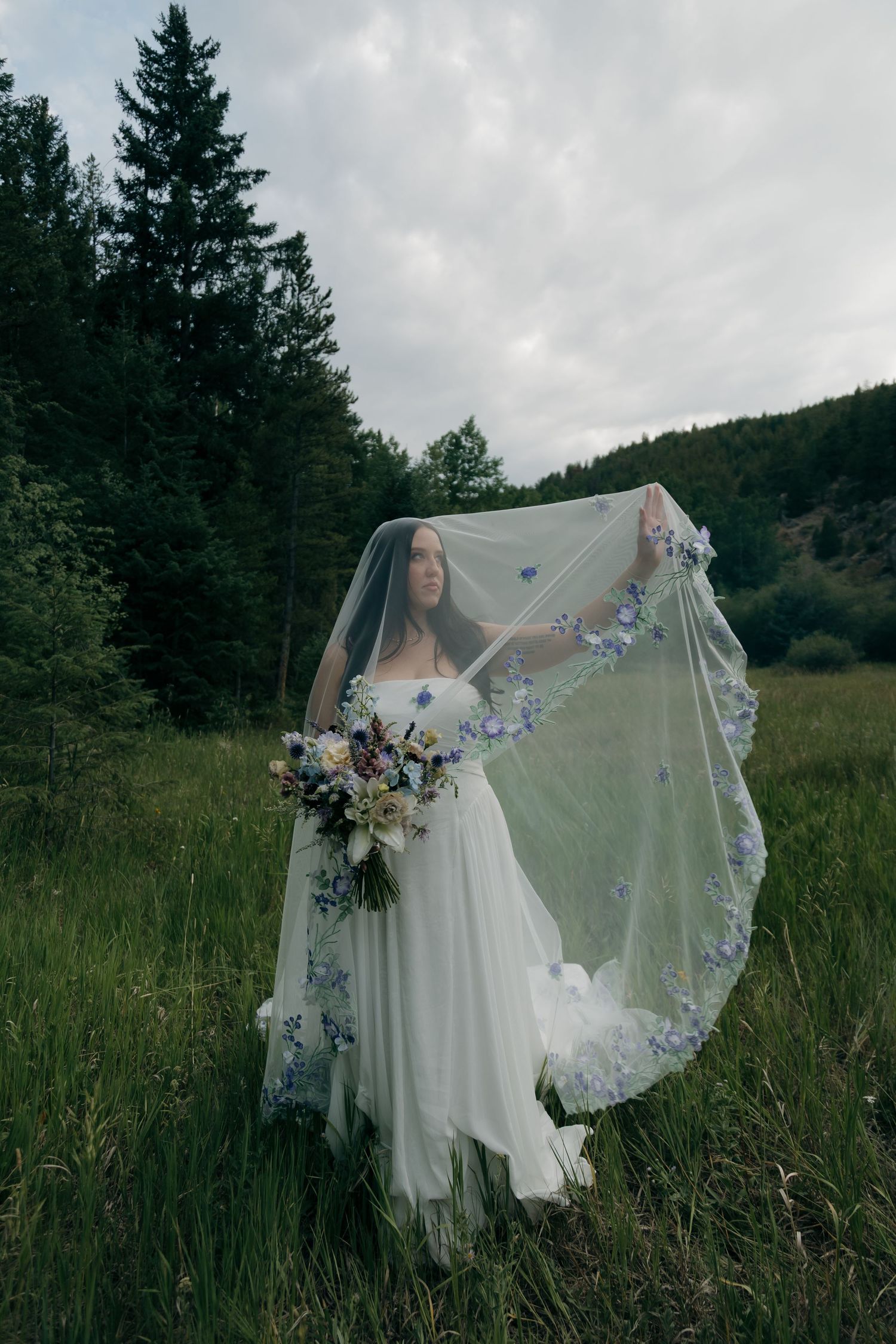 Bride holds her floral-embroidered veil overhead while standing in a mountain meadow, holding a wildflower bouquet in white dress.