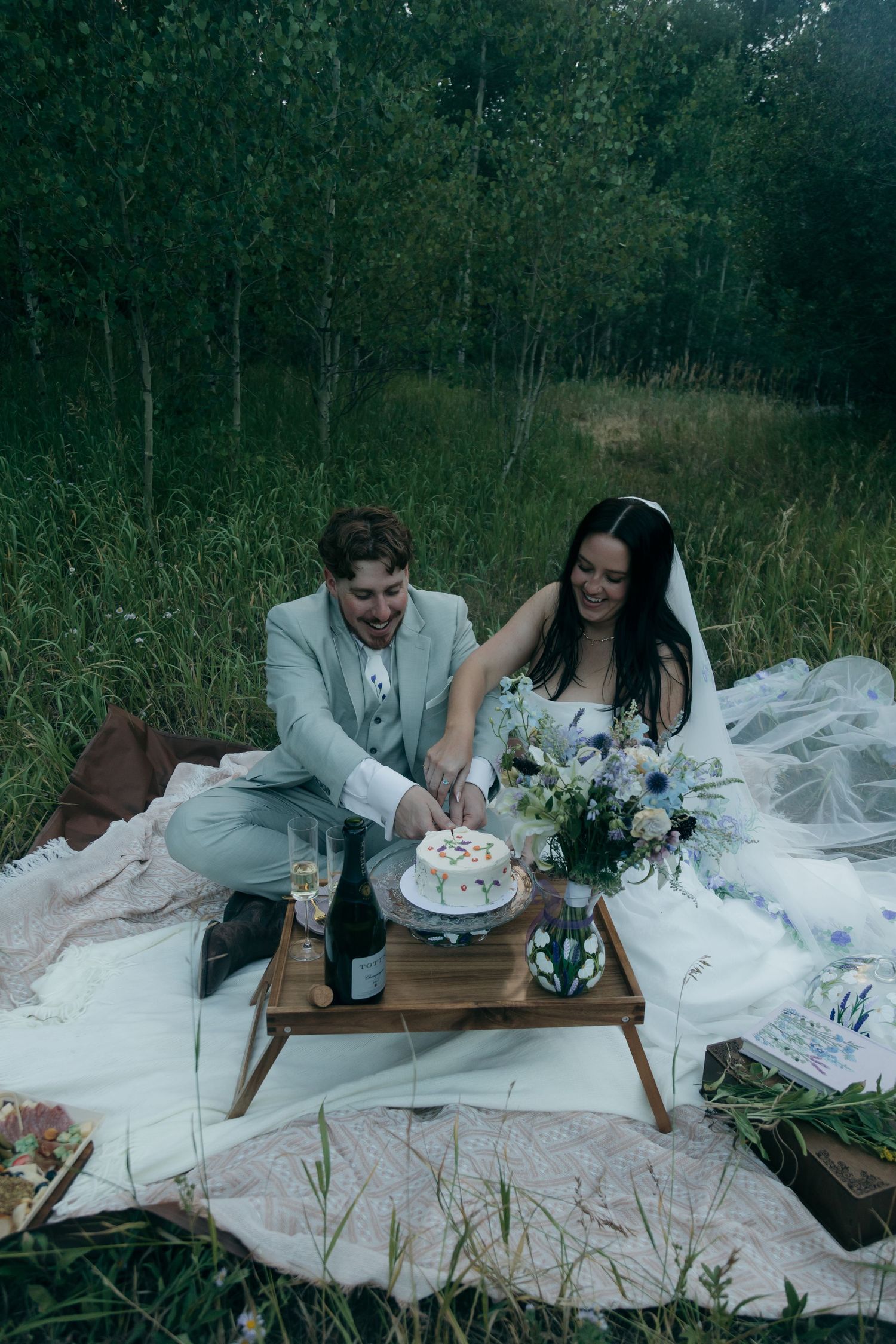 Couple cuts wedding cake together on picnic blanket during intimate forest elopement celebration.