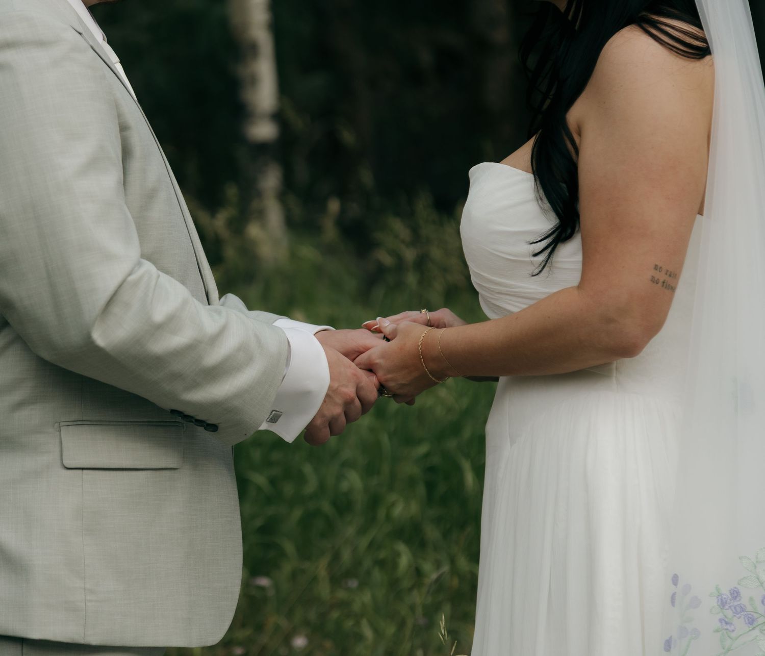 A bride and groom hold hands during their outdoor wedding ceremony, she in a white gown, he in a light gray suit.