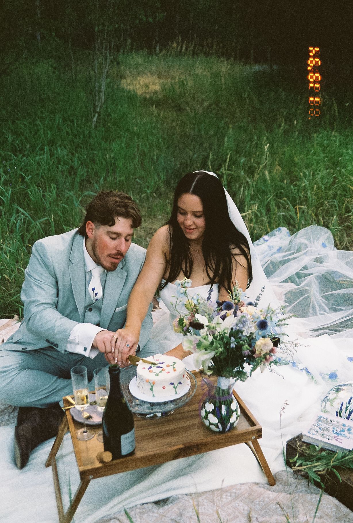 Bride and groom cut a decorated cake together during an outdoor evening picnic with flowers and champagne on film.