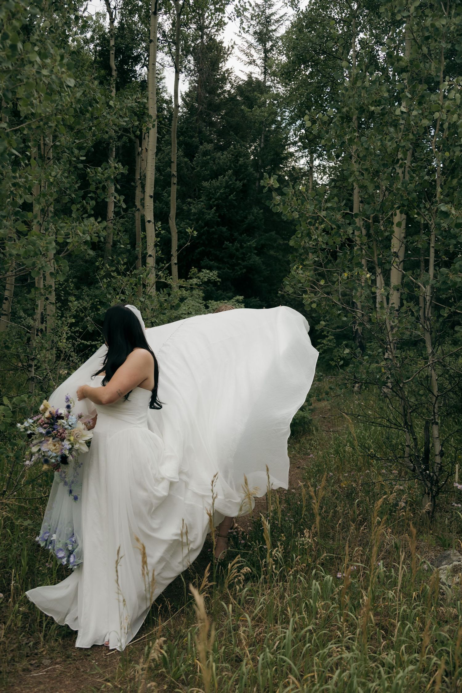 Bride in flowing white gown holding wildflower bouquet walks through lush forest trail on wedding day.