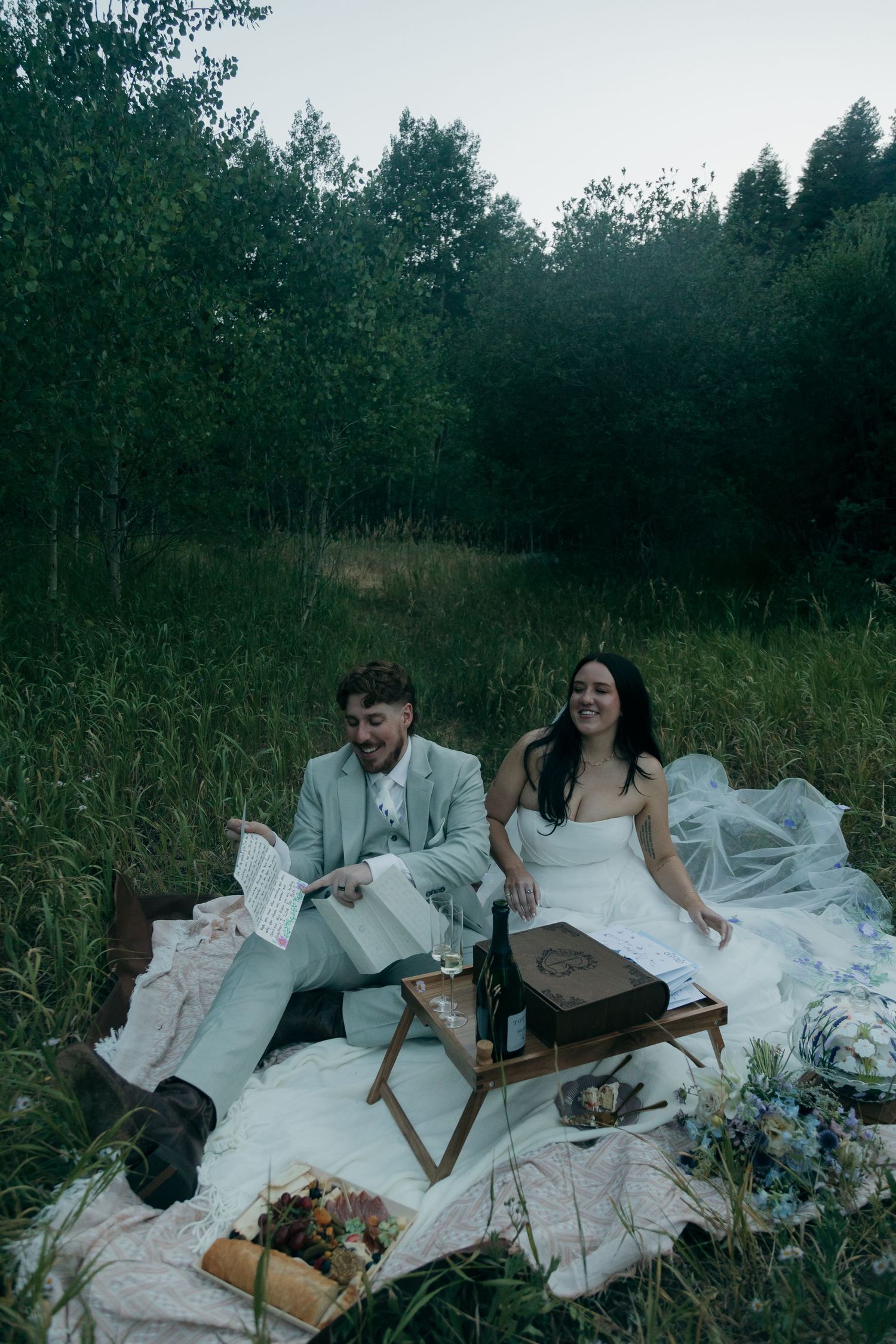 Bride and groom sit on a white picnic blanket in a forest meadow reading letters at their elopement.