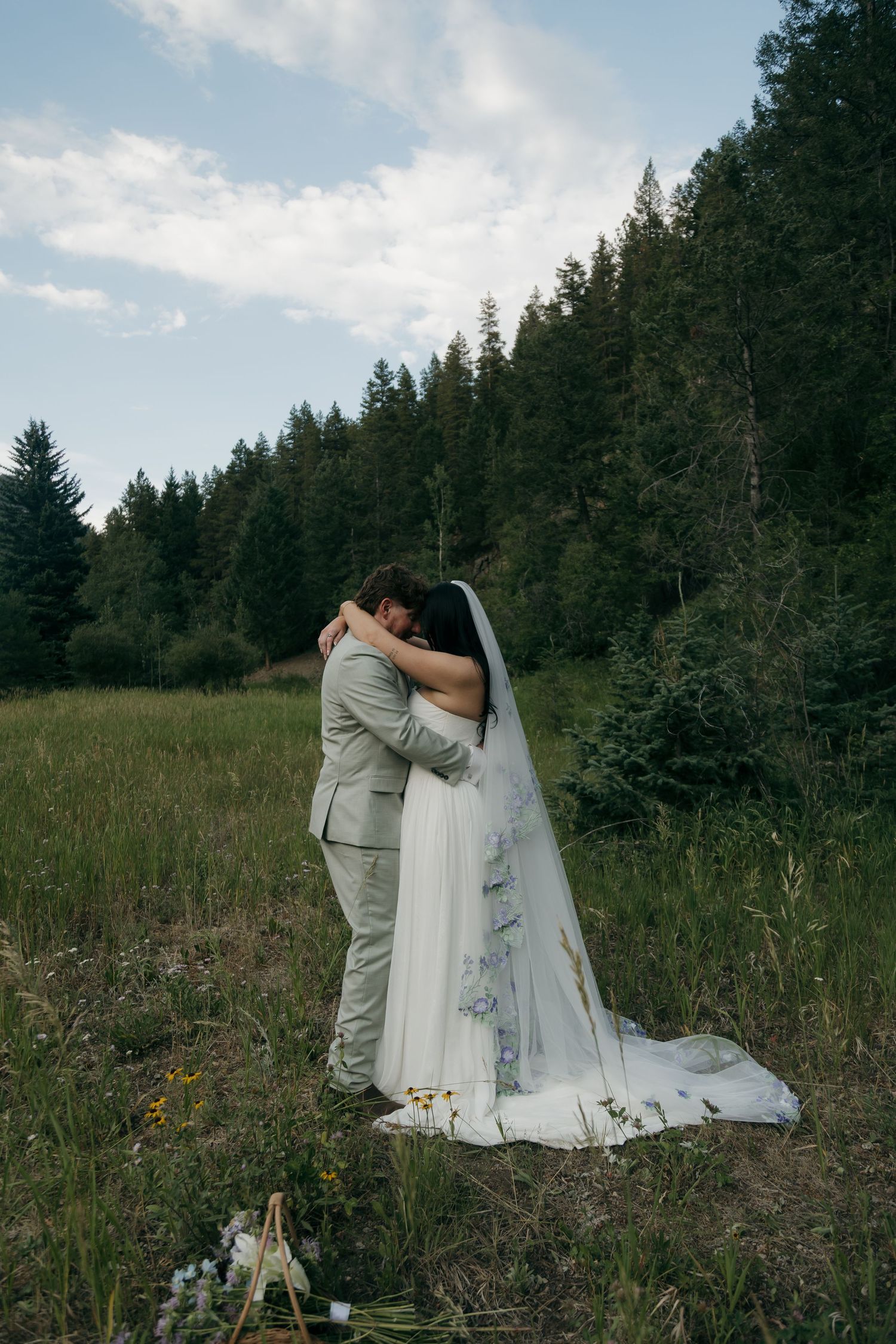 Newlyweds embrace in a sweeping green meadow with forest backdrop and floral bouquet nearby.