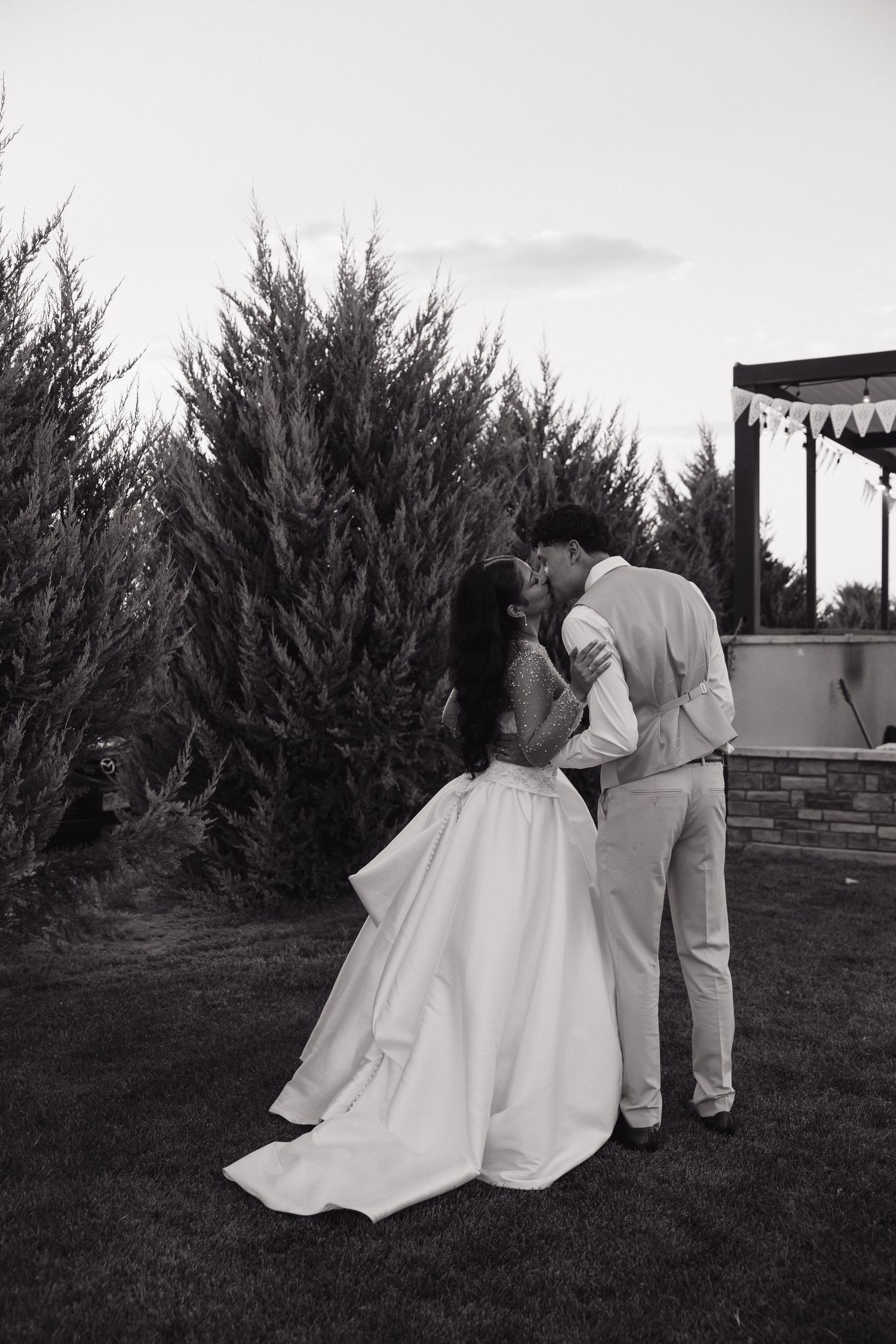 Black and white portrait of bride and groom sharing kiss in garden at dusk.