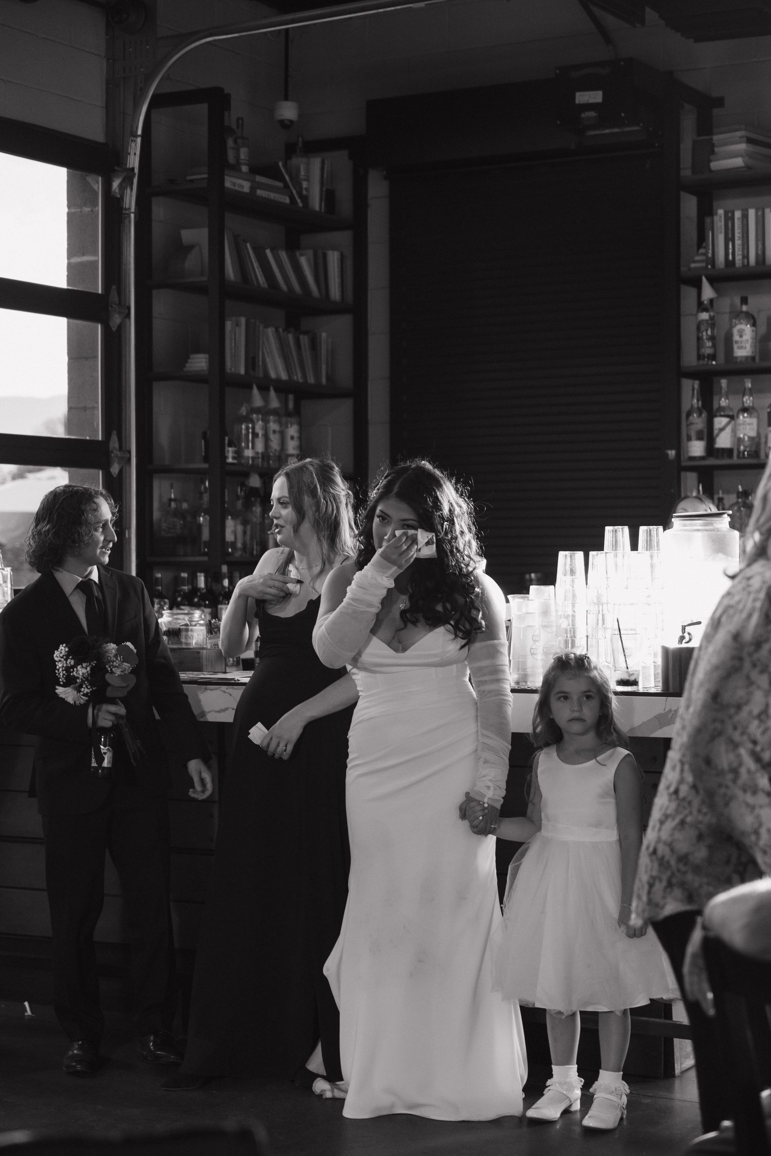 Black and white photo of bride in white gown with bridesmaids and flower girl at indoor bar.