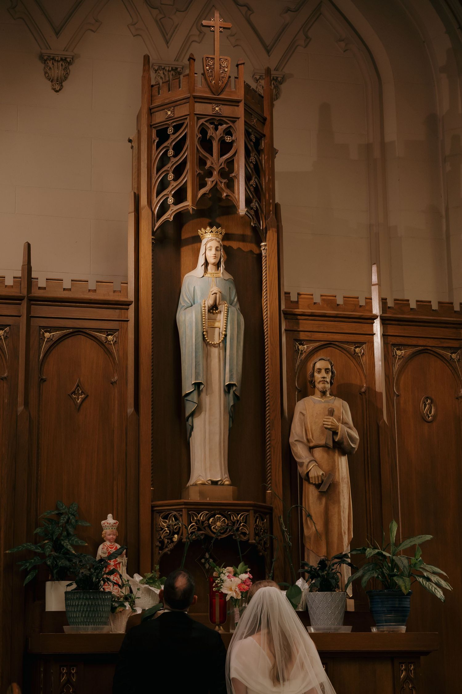 Couple kneeling before Virgin Mary statue at ornate Catholic church altar during wedding ceremony.
