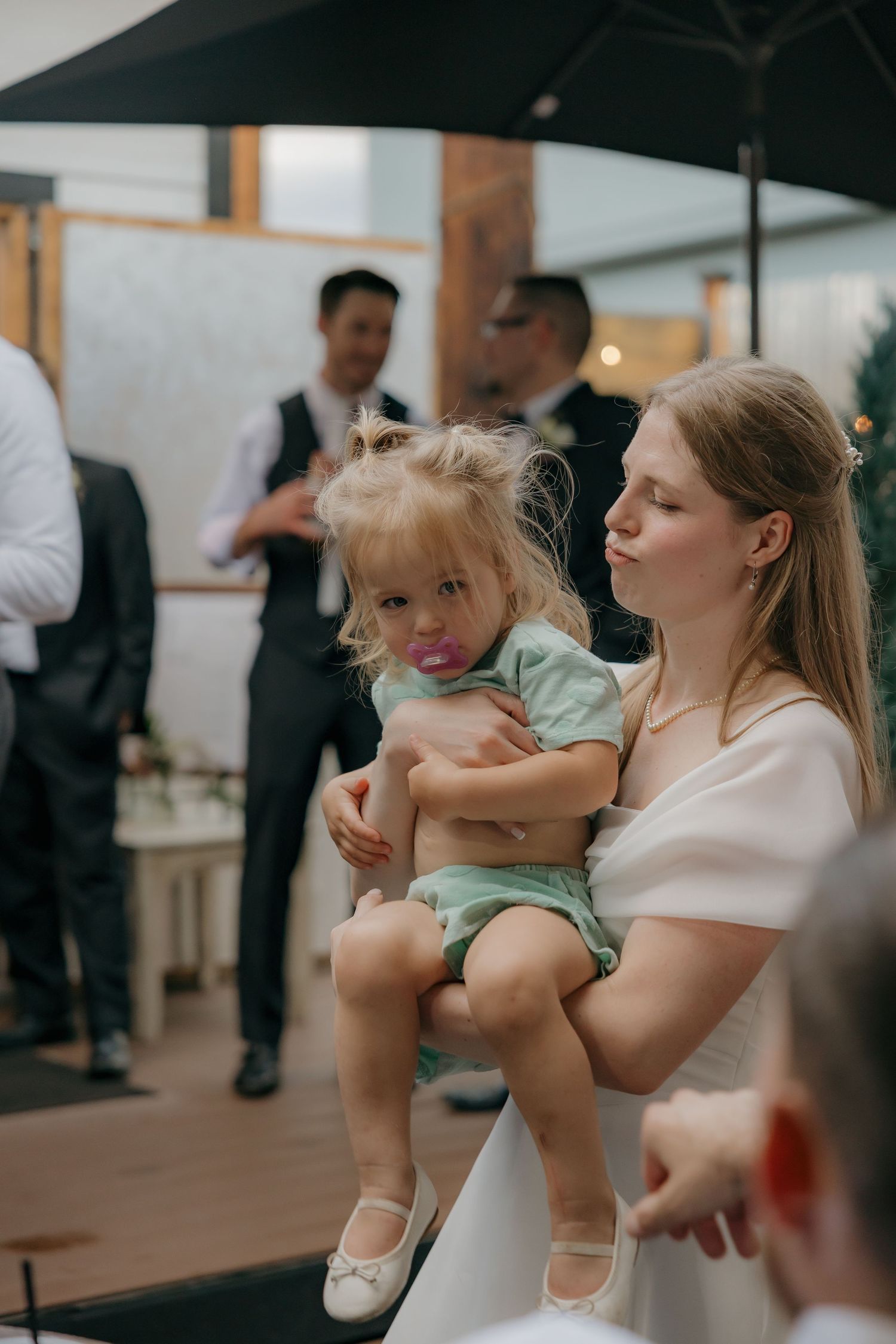 Young mother in white dress holding toddler girl with pacifier at elegant wedding reception.