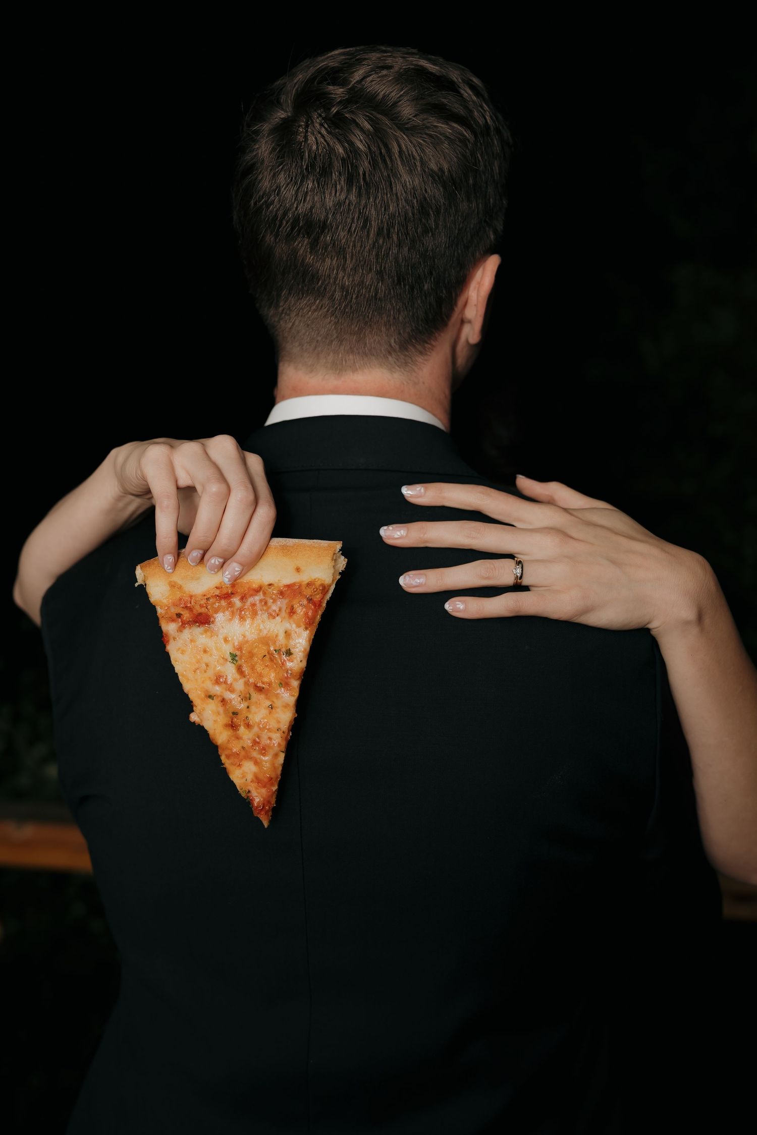 Bride hugs groom from behind holding pizza slice against his black suit jacket at night.