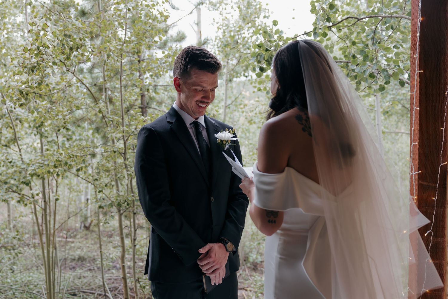 Groom smiling emotionally as bride reads vows during intimate outdoor wedding ceremony among aspen trees.