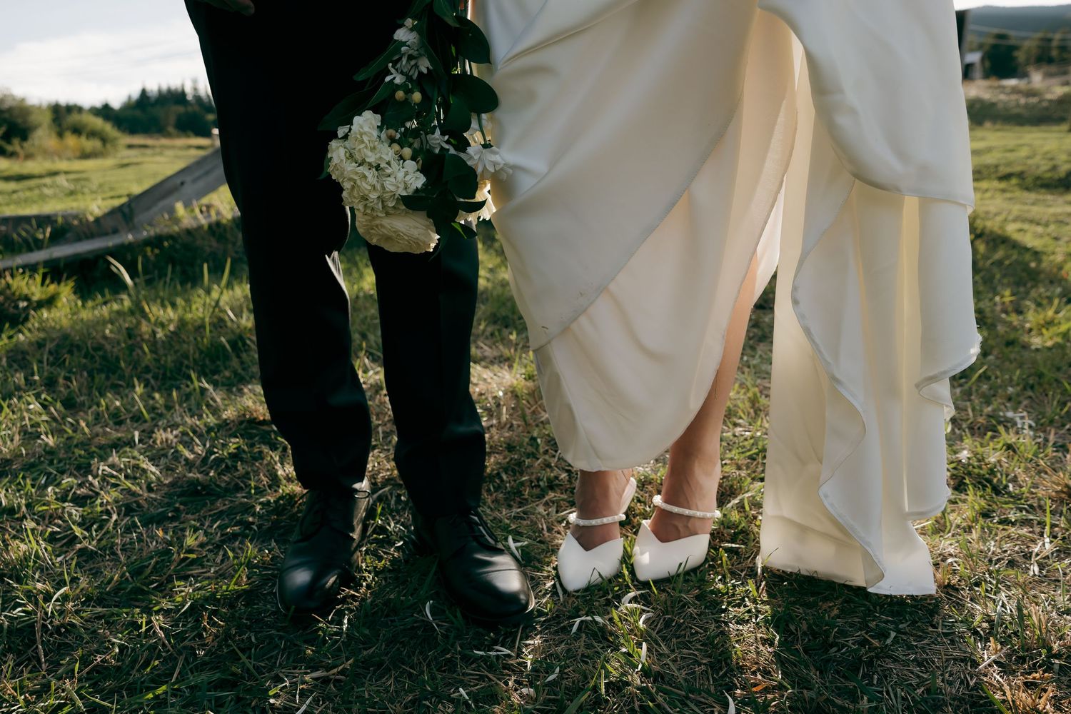 Close-up of bride and groom's feet on grass, bride wearing white heels at outdoor wedding.