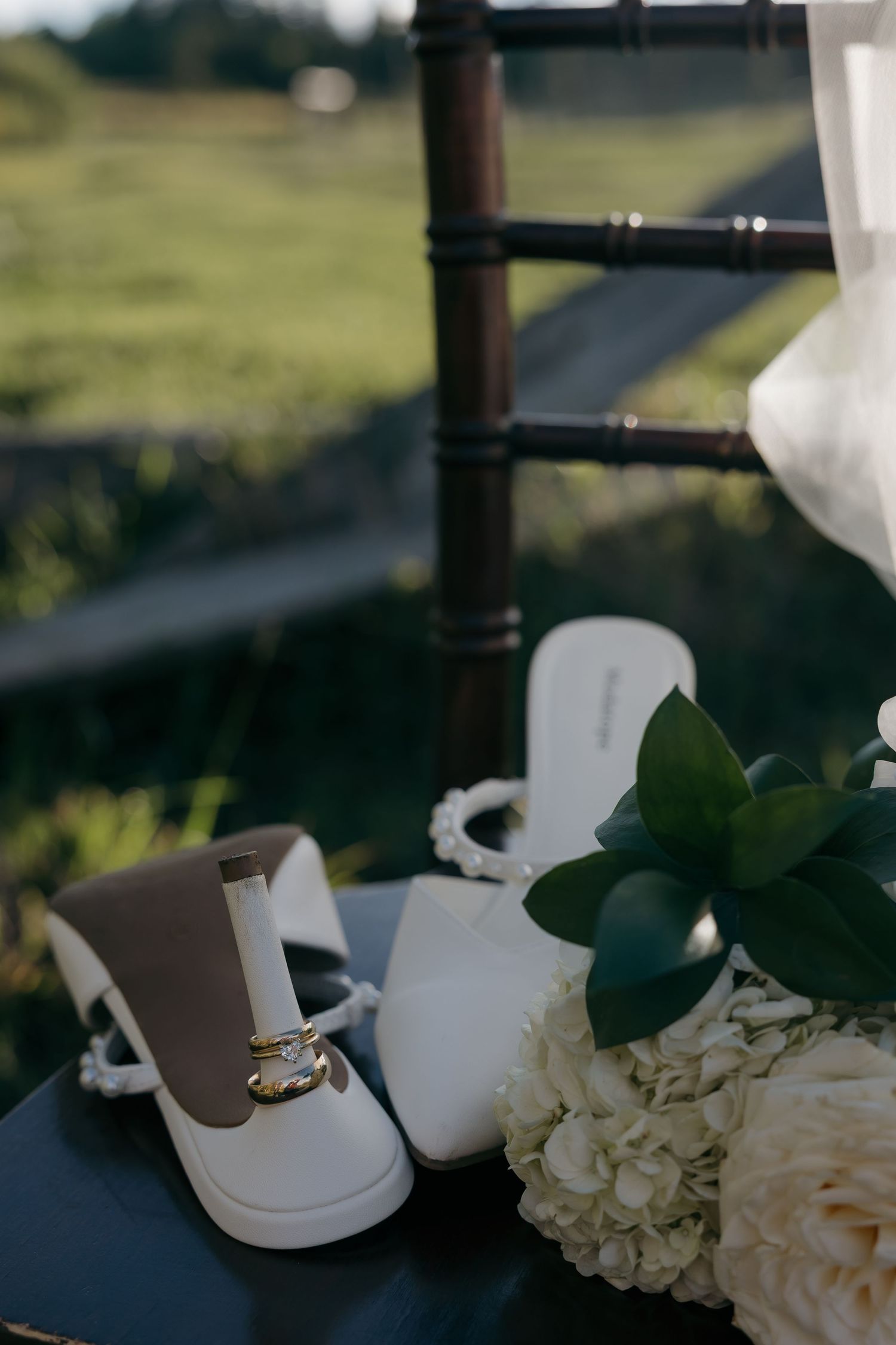 White bridal heels with gold wedding rings resting on shoe near white flowers.