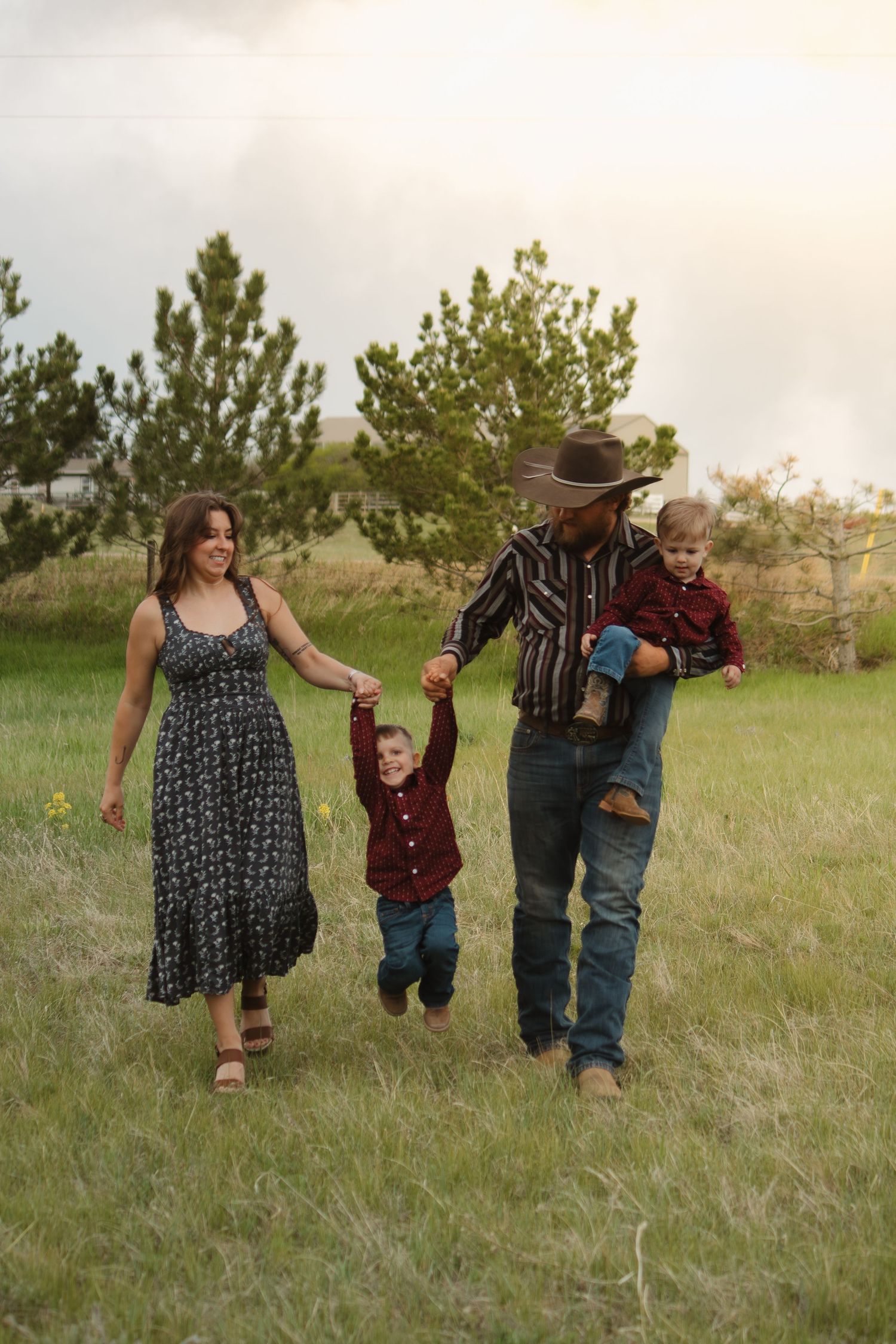 A family walks hand in hand through a meadow with pine trees in the background at sunset.