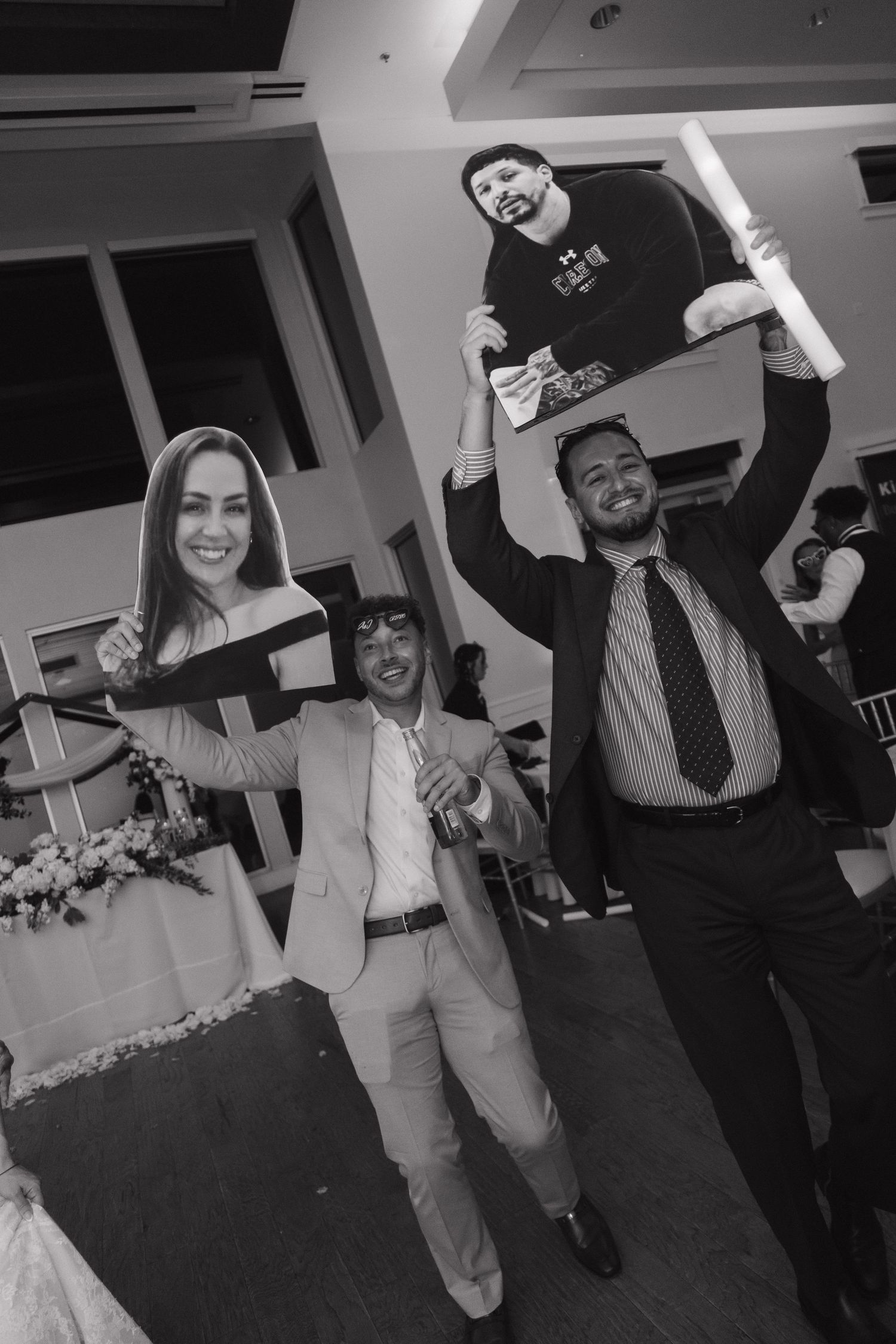 Two men hold up cardboard cutouts of bride and groom at wedding reception, black and white photo.