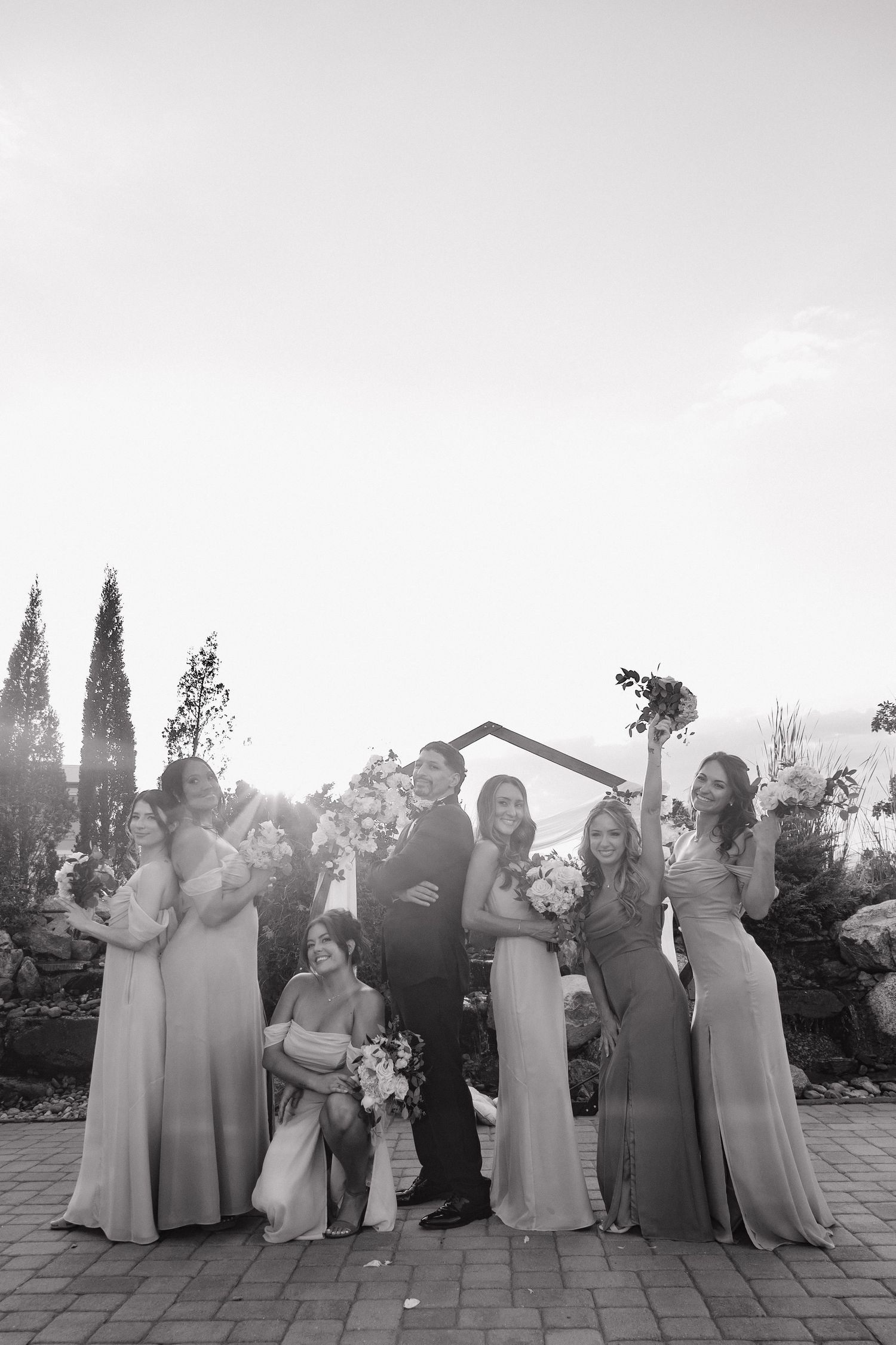 Bride and groom pose with bridesmaids outdoors at sunset in elegant black and white wedding photo.