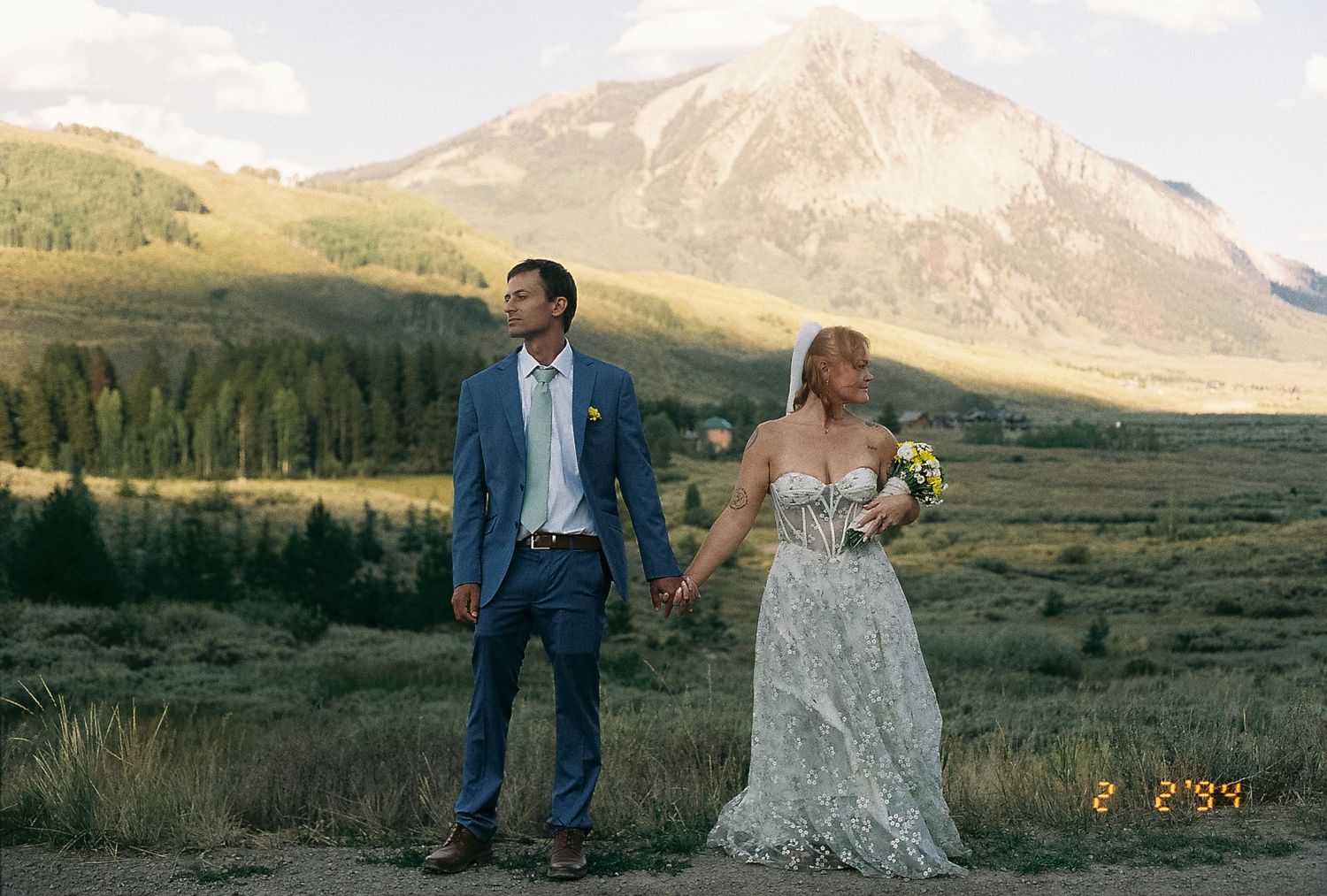 Bride and groom holding hands in wildflower meadow with dramatic mountain peak backdrop at golden hour.