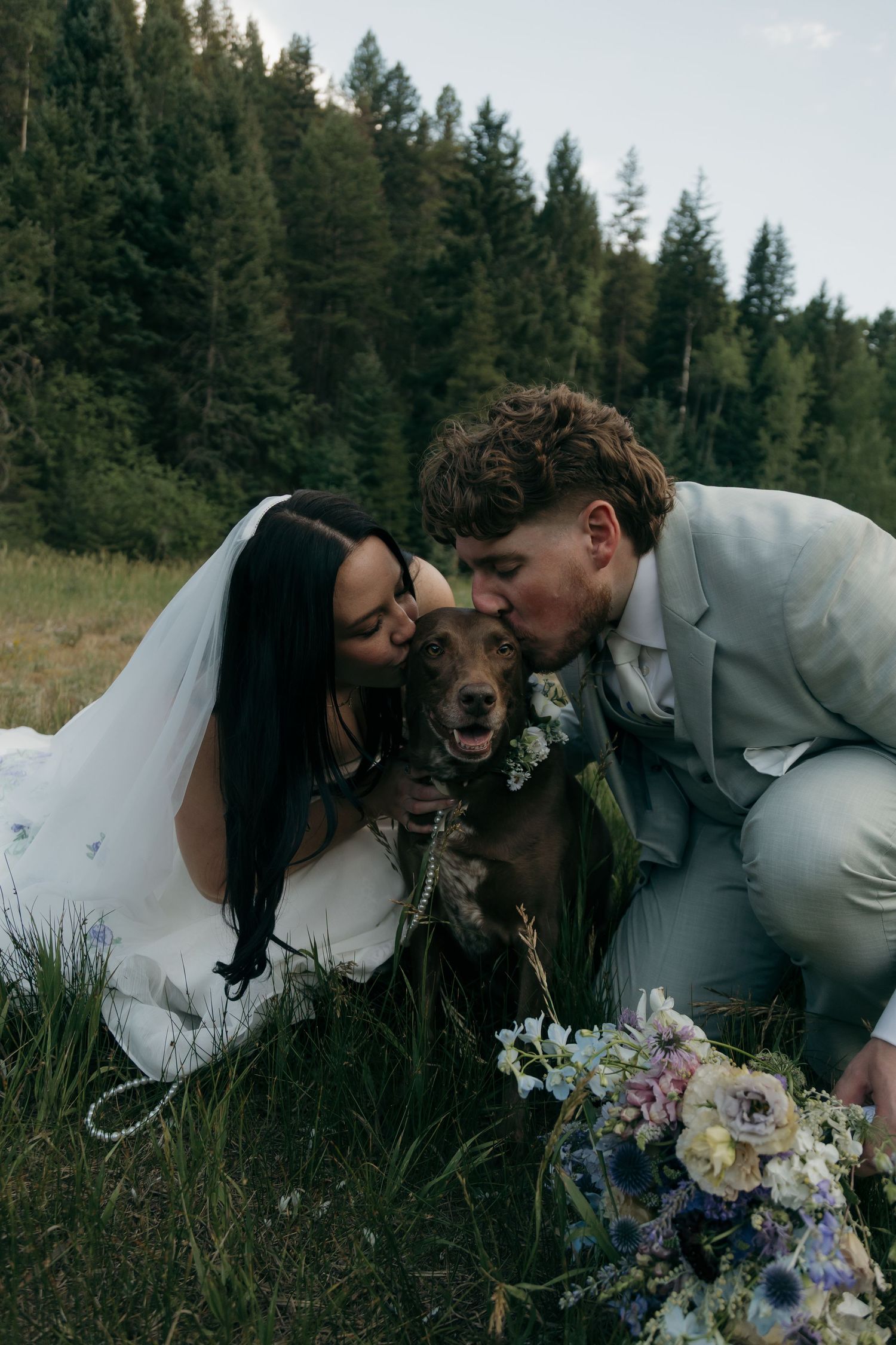 Bride and groom kiss their brown dog wearing a floral collar during outdoor forest wedding celebration.