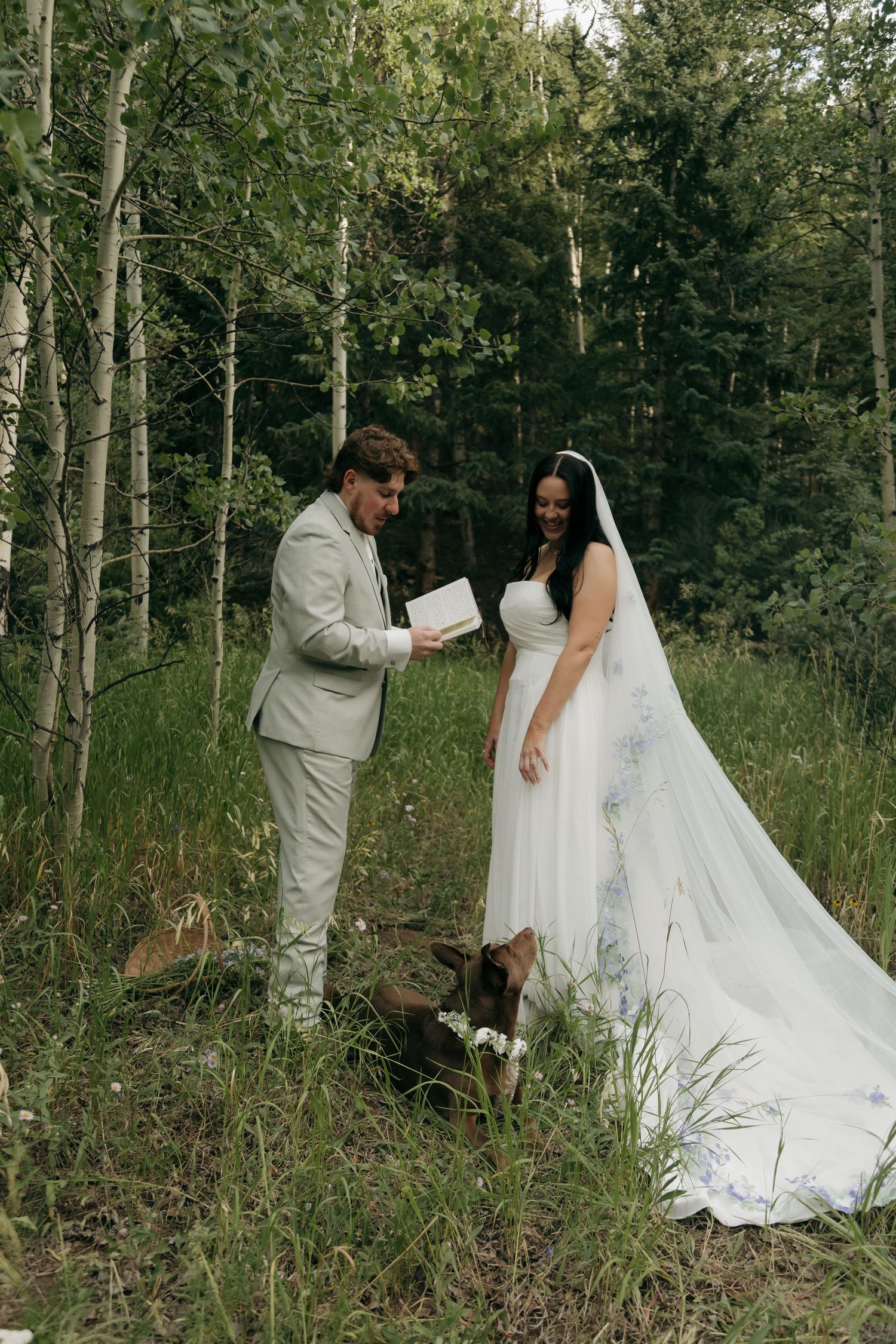 Bride and groom share vows in a lush forest setting with their chocolate Labrador dog beside them.