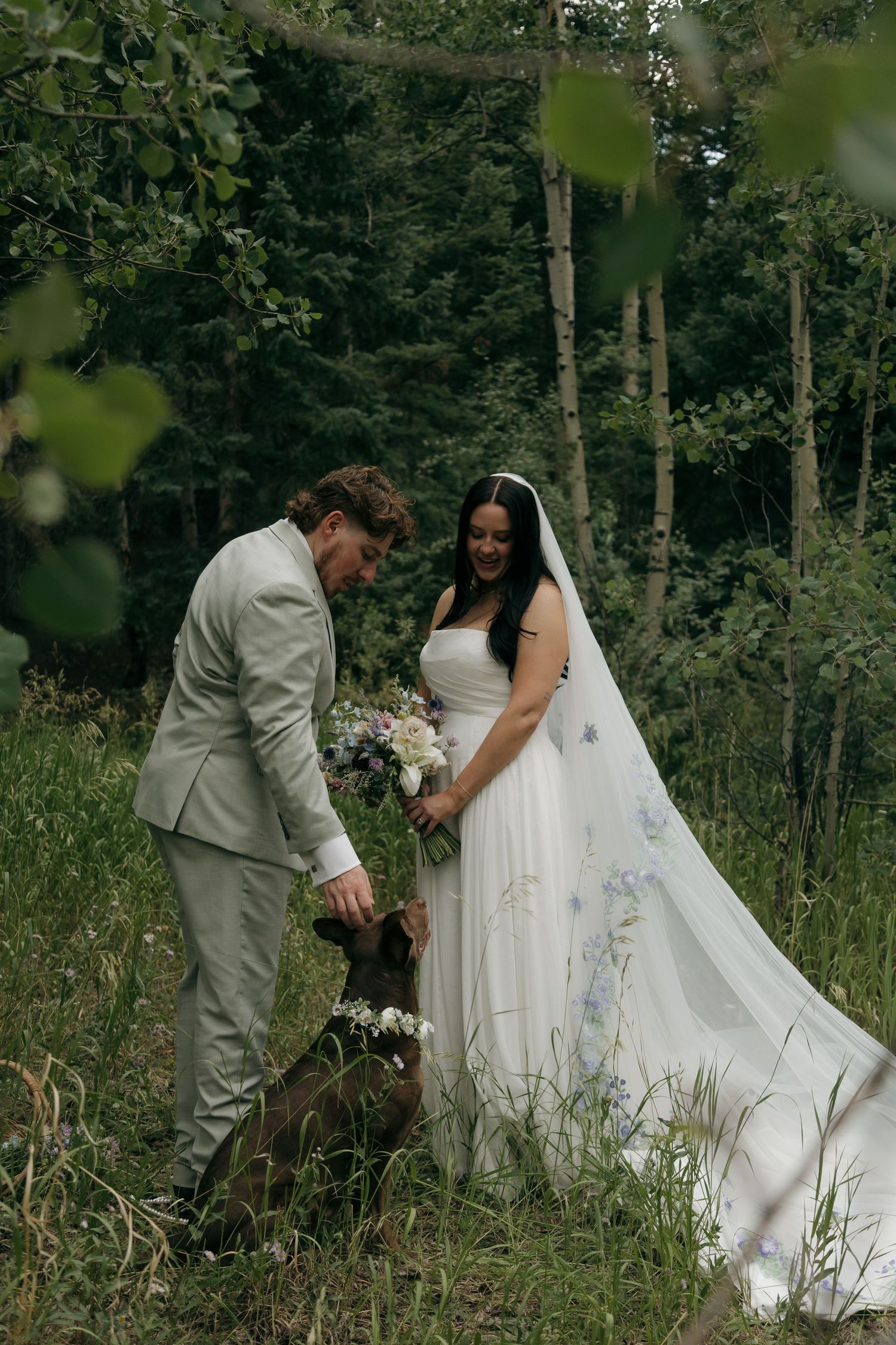 Bride and groom petting their dog in a lush forest setting during their outdoor wedding ceremony.