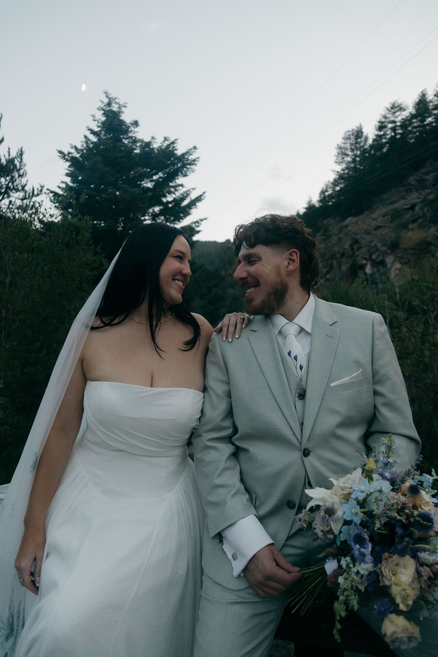 Smiling bride in strapless white gown and groom in light gray suit hold a blue floral bouquet during their outdoor wedding.