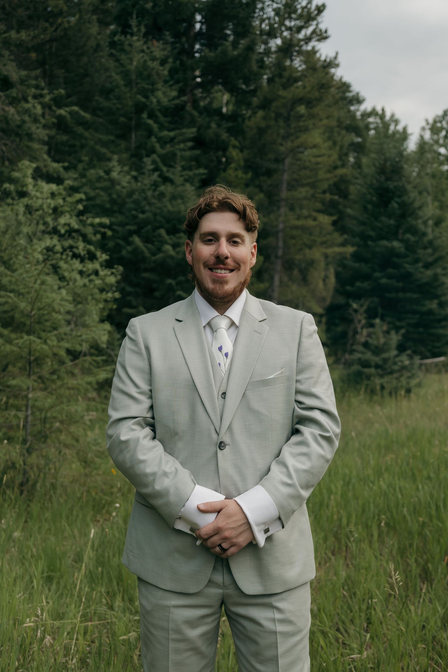 Handsome groom in sage green suit smiling confidently in a green forest meadow on his wedding day.