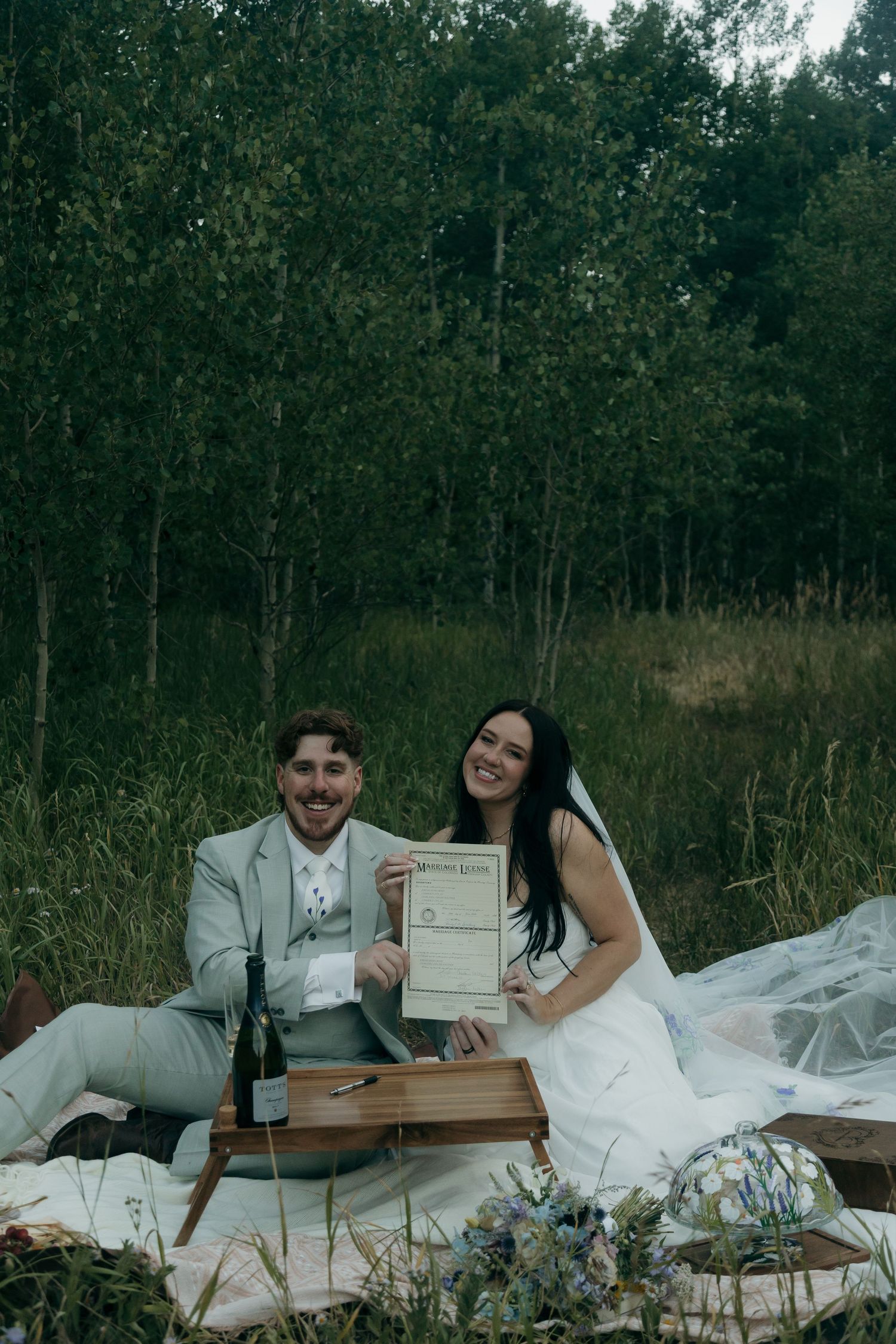 Newlywed couple sitting on a blanket outdoors holding their marriage certificate, dressed in wedding attire near a forest.