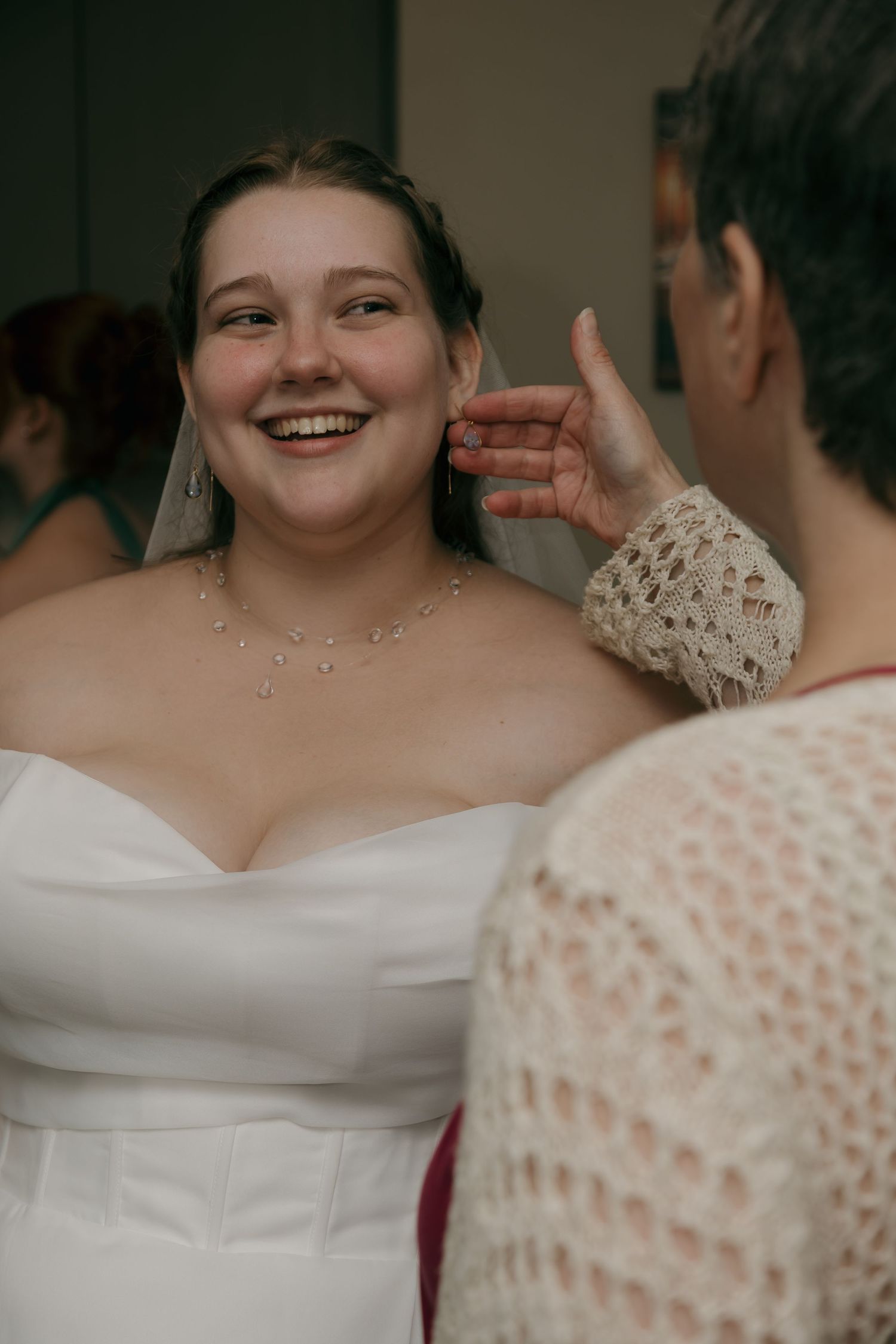 Smiling bride in white strapless gown wearing veil and crystal necklace during wedding preparation.