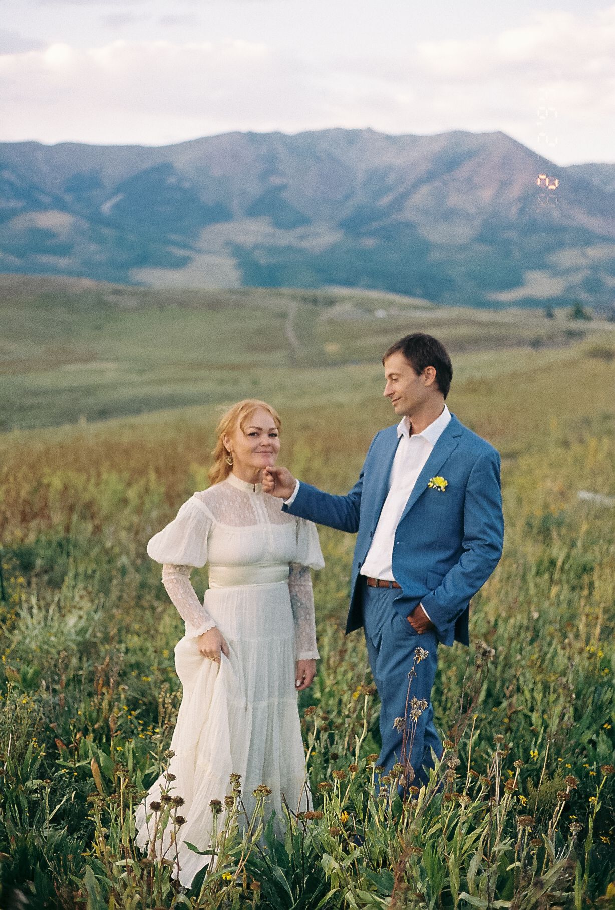 Couple in wedding attire standing in a lush mountain meadow, bride in white dress and groom in blue suit.