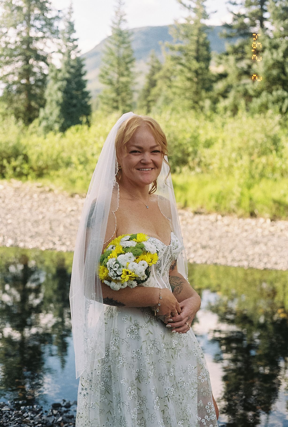 Smiling bride in lace wedding dress holds yellow bouquet by mountain stream with pine trees.