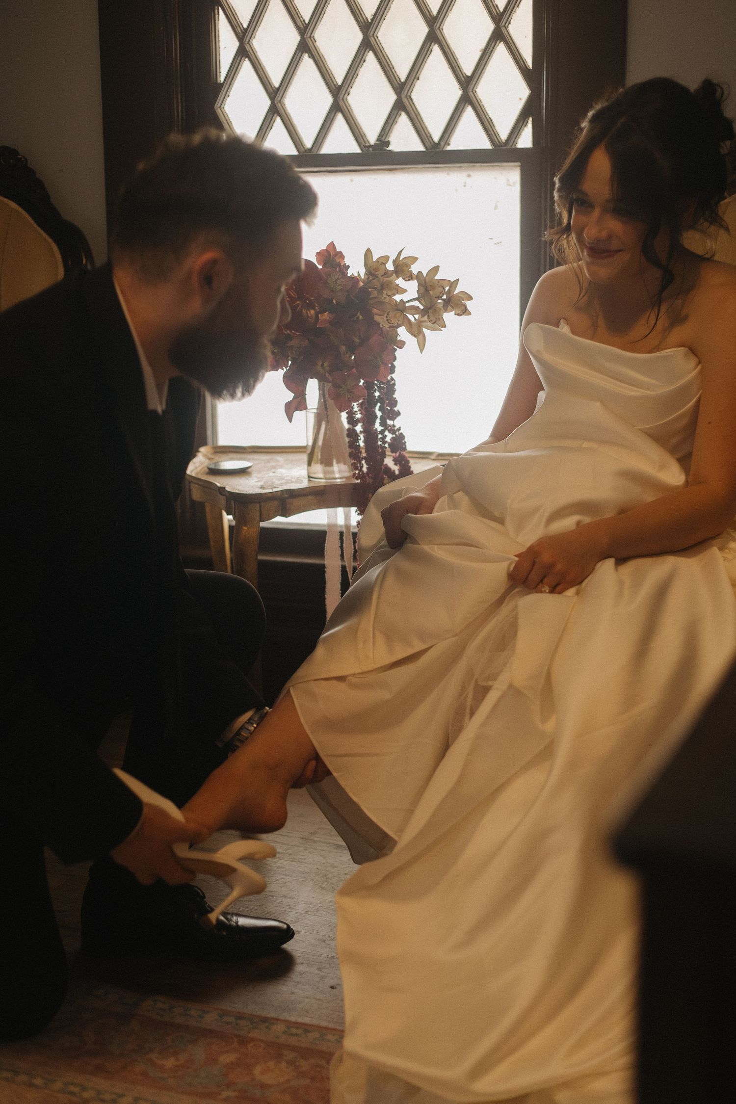 A wedding dress fitting scene shows the bride getting her shoes adjusted near a window with flowers.