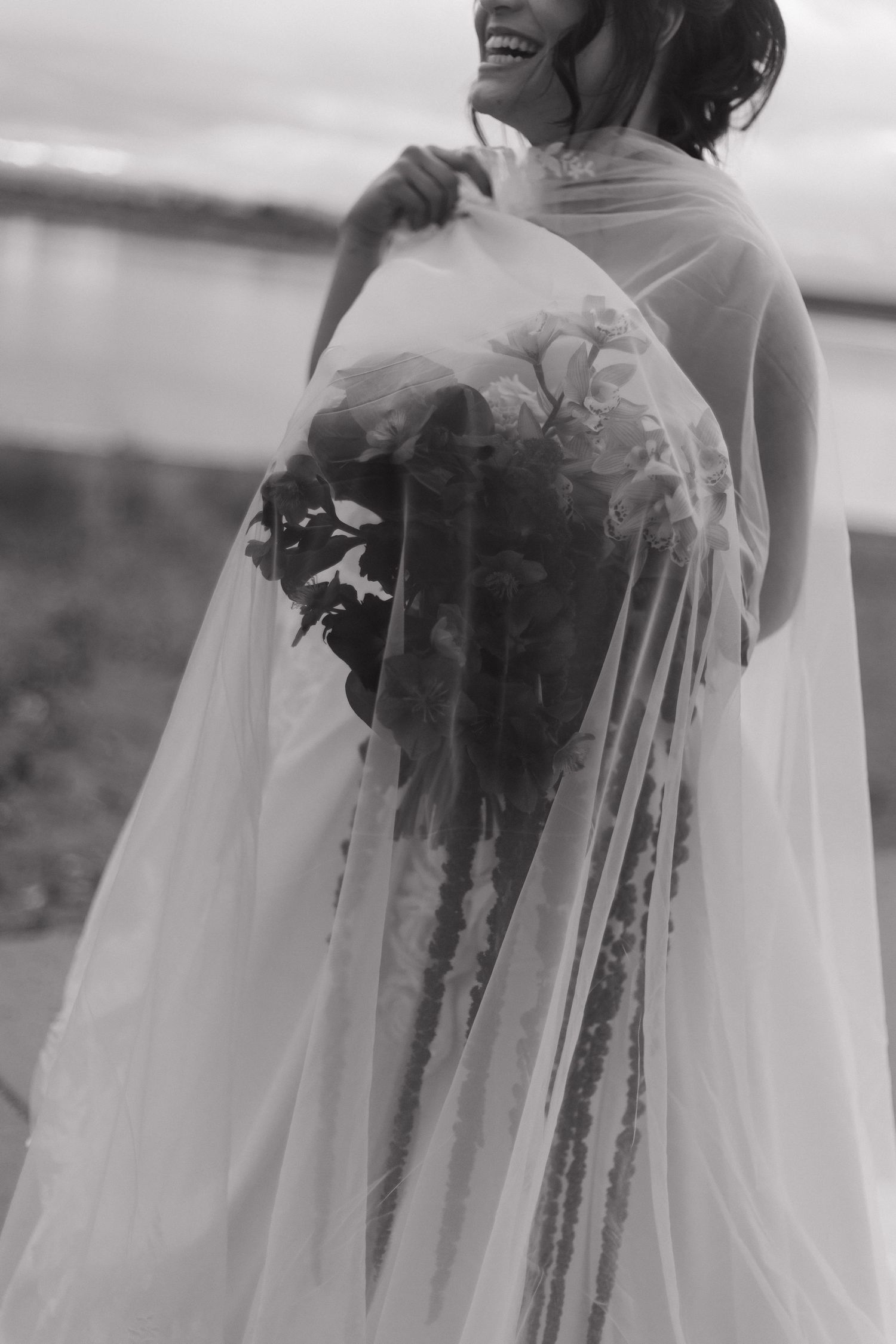 A bride in a flowing veil holds a cascading bouquet against a scenic backdrop.