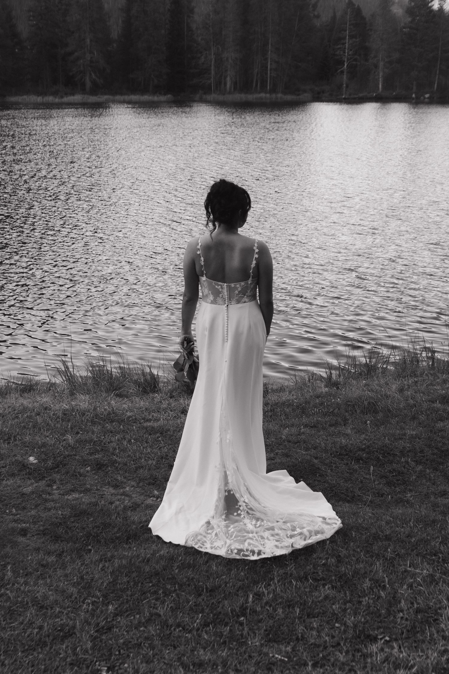 A bride in a white dress stands alone by a tranquil lake in black and white.