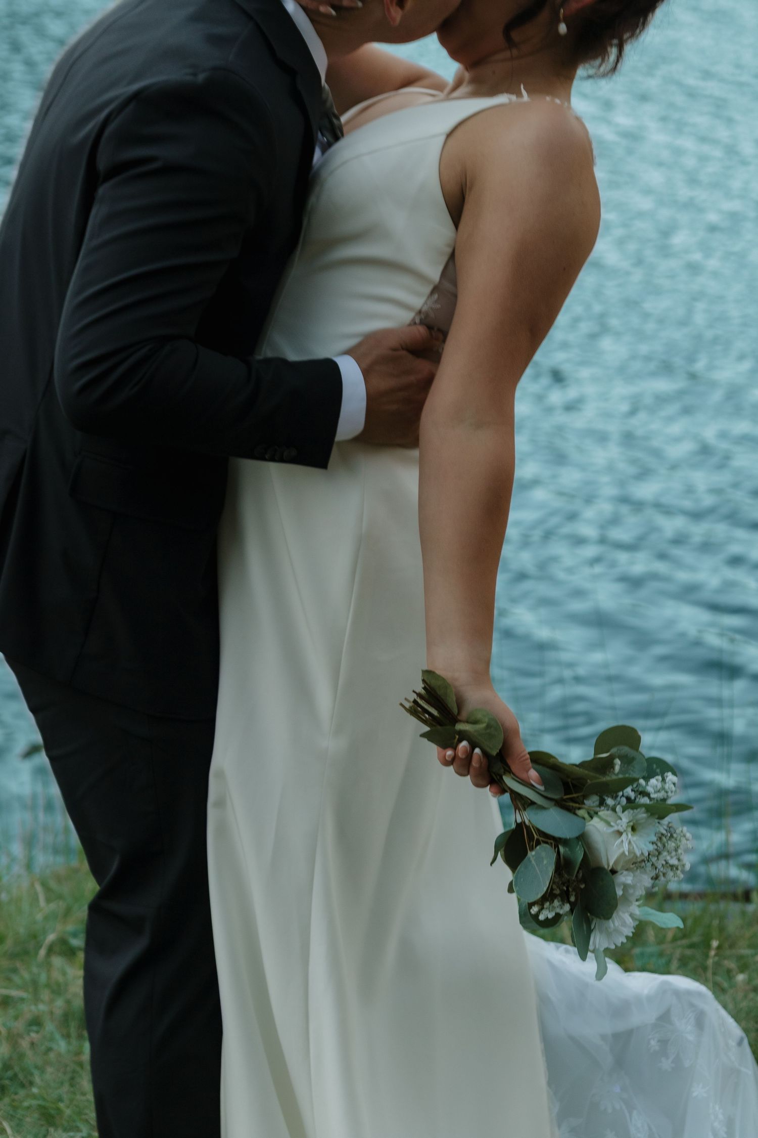 Close-up of newlyweds embracing by a lake with the bride holding a bouquet.