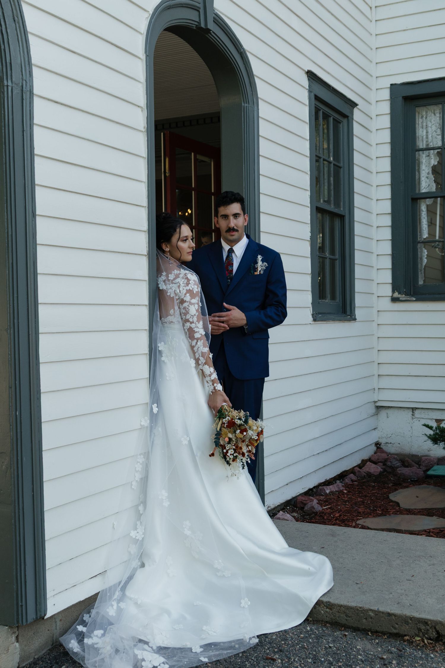 Couple poses for portraits outside white building with grey trim and arched windows.