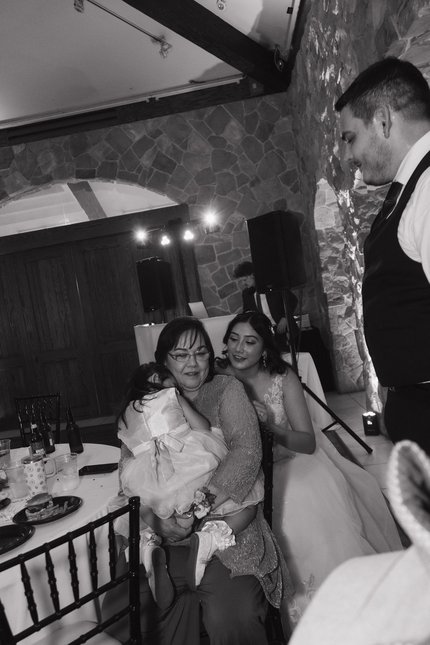 Bride embraces seated elderly woman while groom looks on at wedding reception in stone venue.