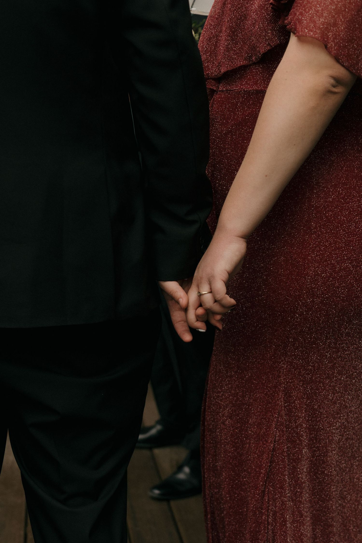 Close-up of couple holding hands, woman in sparkling burgundy dress and man in black suit at wedding.