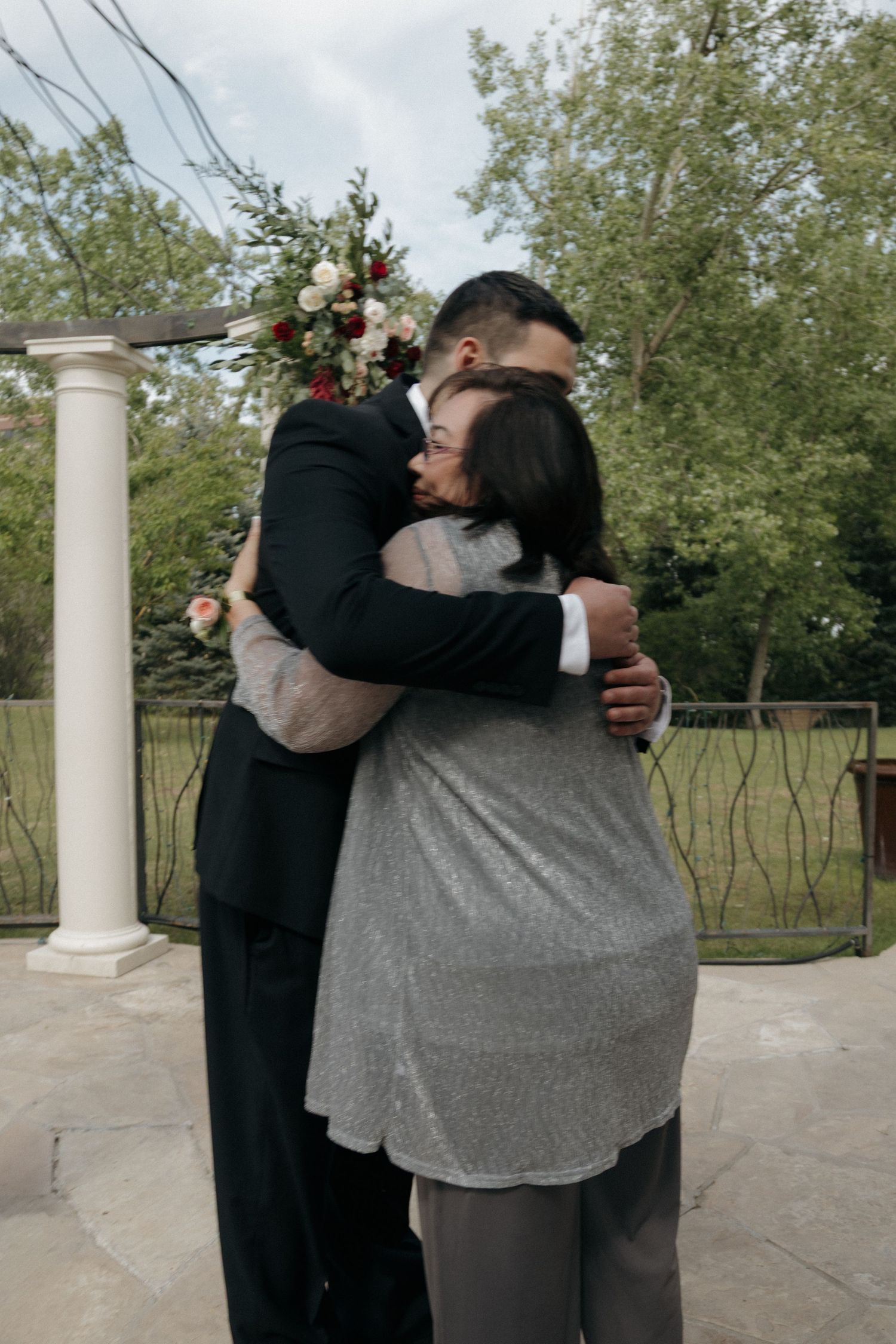 Groom in black suit shares a warm embrace with a female guest near a floral wedding arch.