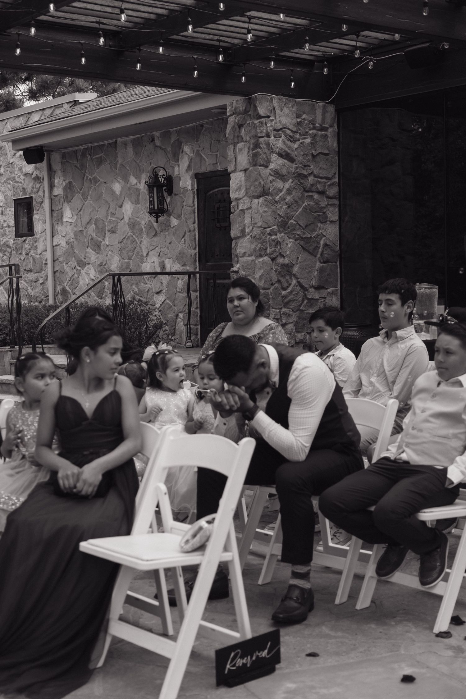 Black and white photo of wedding guests seated outdoors during a ceremony at a stone venue with string lights.