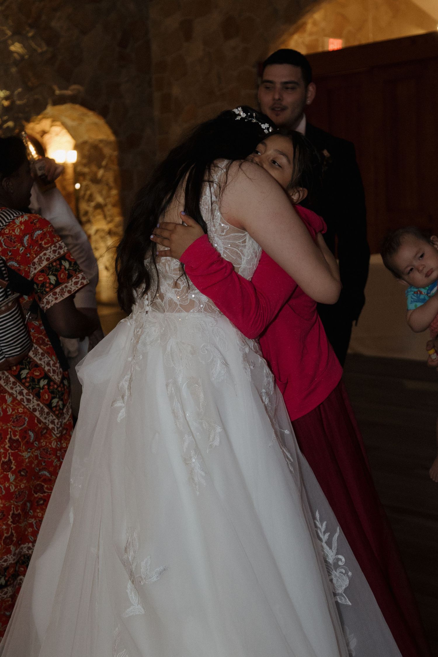 Bride in lace wedding gown sharing a heartfelt hug with a guest in red at a rustic stone wedding venue.