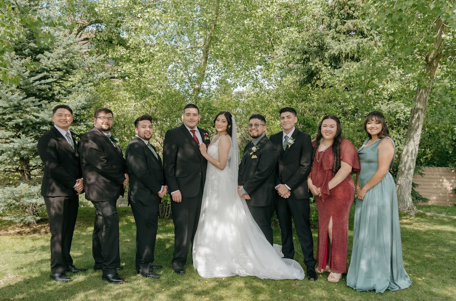 Wedding party group portrait with bride and groom surrounded by lush green trees.