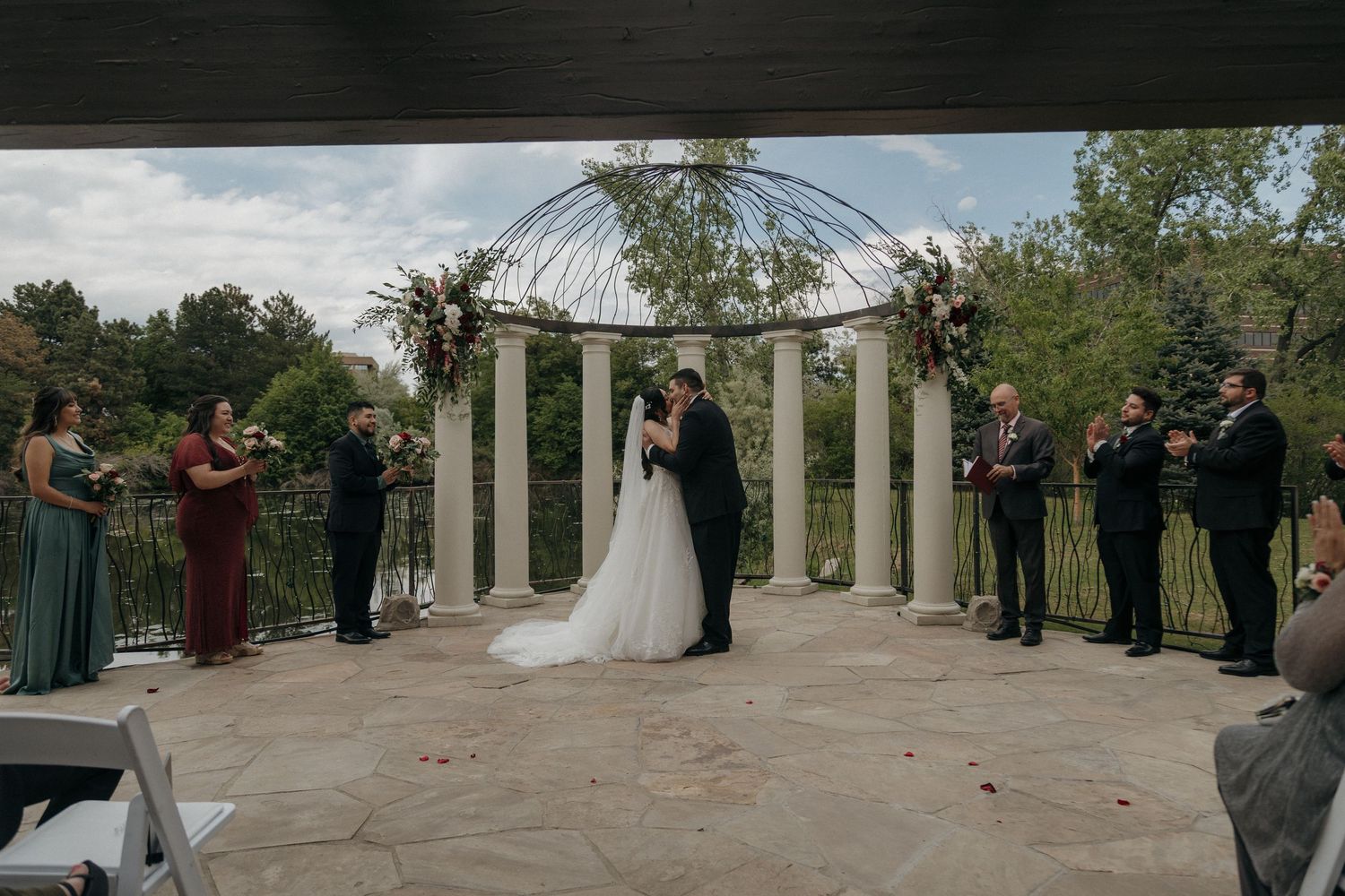 Bride and groom share first kiss at outdoor wedding ceremony surrounded by wedding party beneath floral arch.
