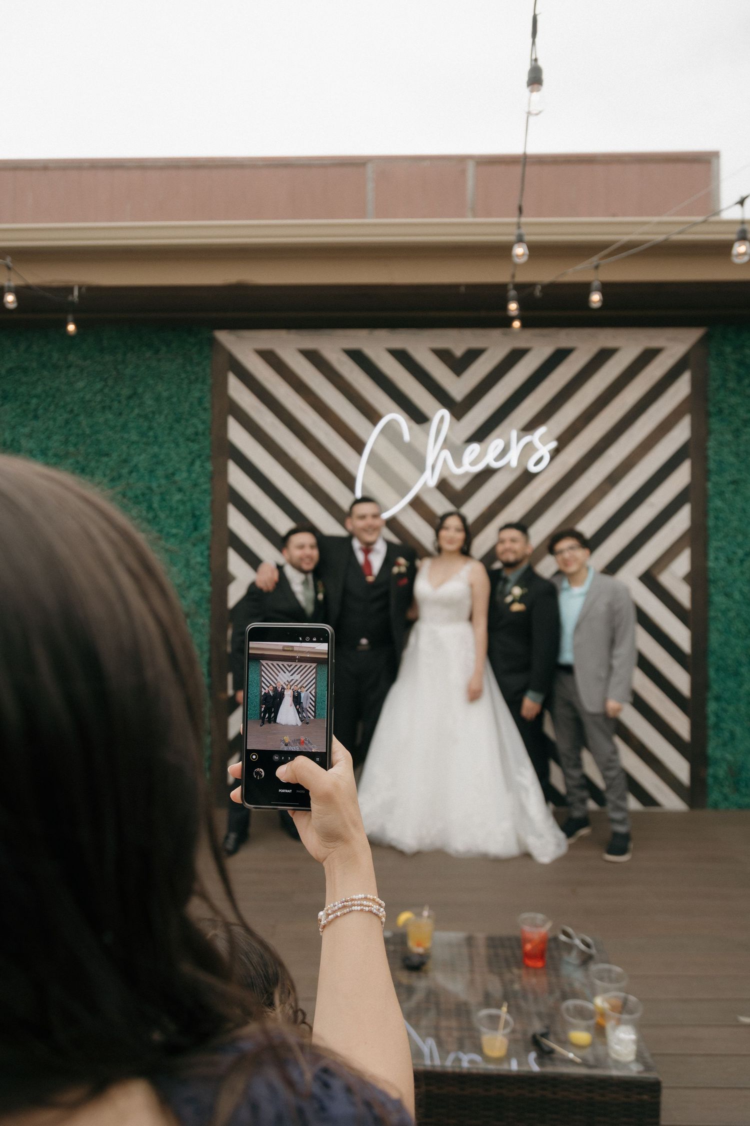 Wedding guests pose for a photo in front of a Cheers neon sign backdrop.