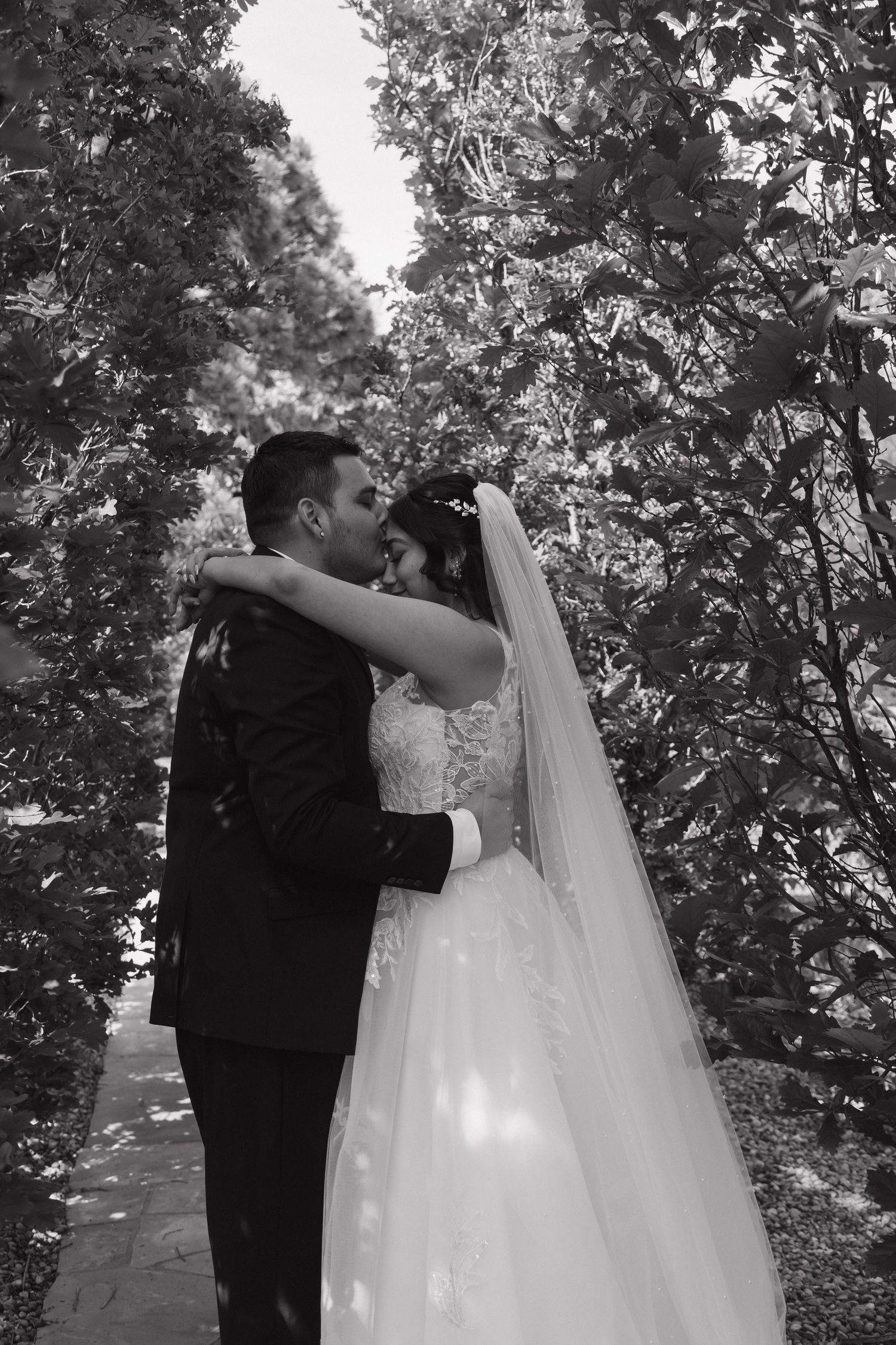 Black and white romantic portrait of bride and groom embracing and kissing among lush trees outdoors.