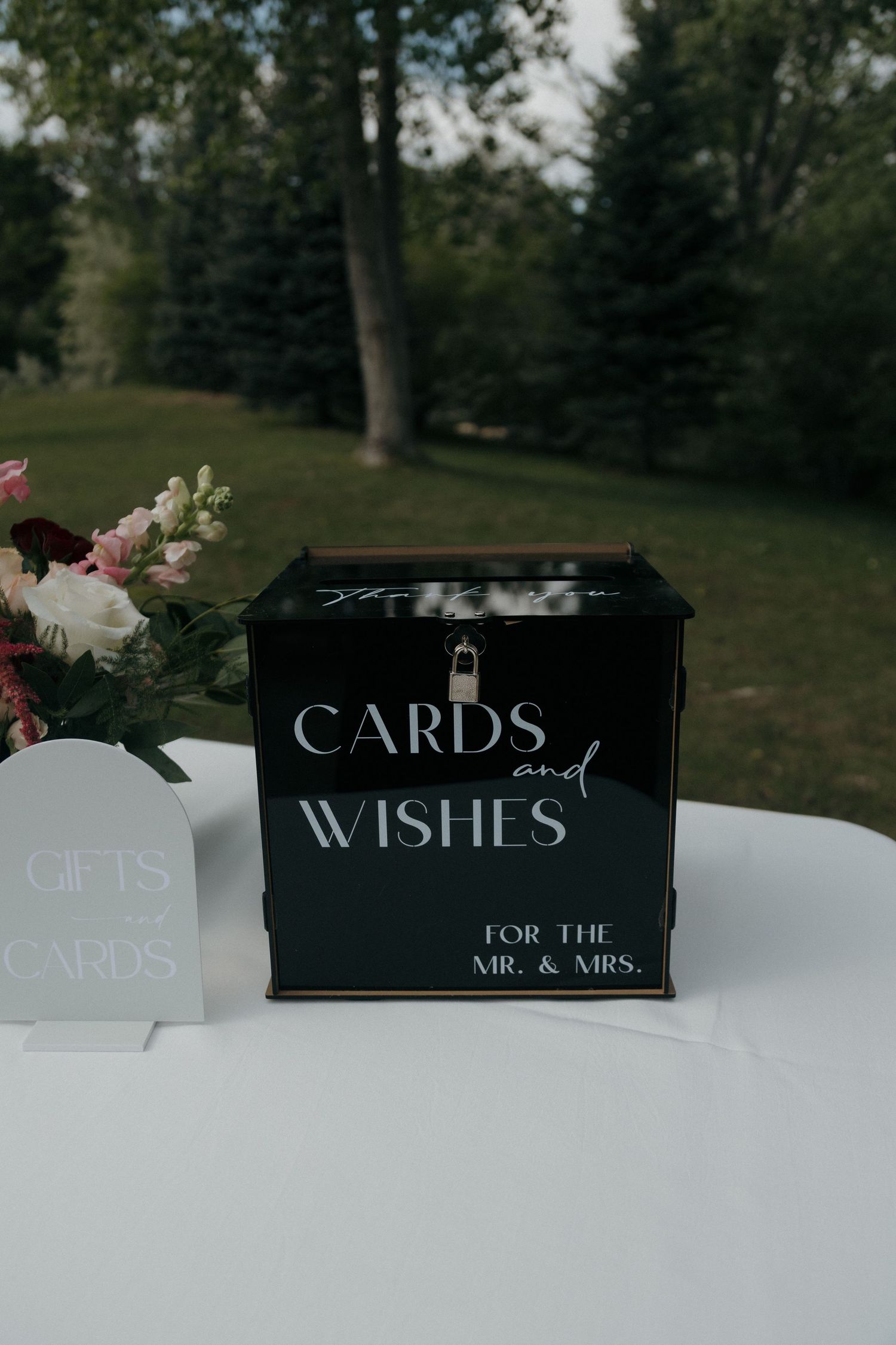 Black wedding card box labeled Cards and Wishes for the Mr. and Mrs. displayed on outdoor table with floral arrangement.