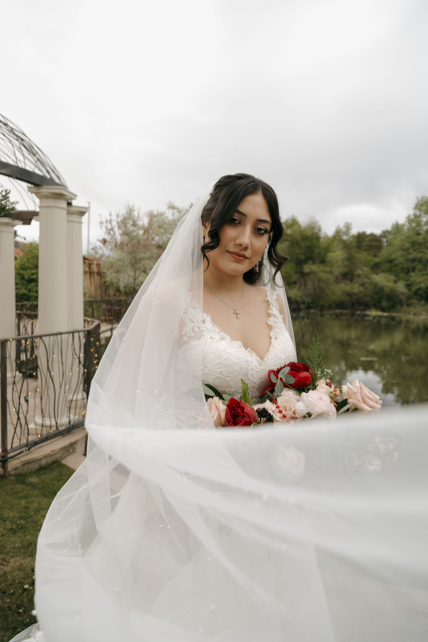 Bride in lace wedding gown holding a red and pink bouquet with veil flowing near a lakeside gazebo.