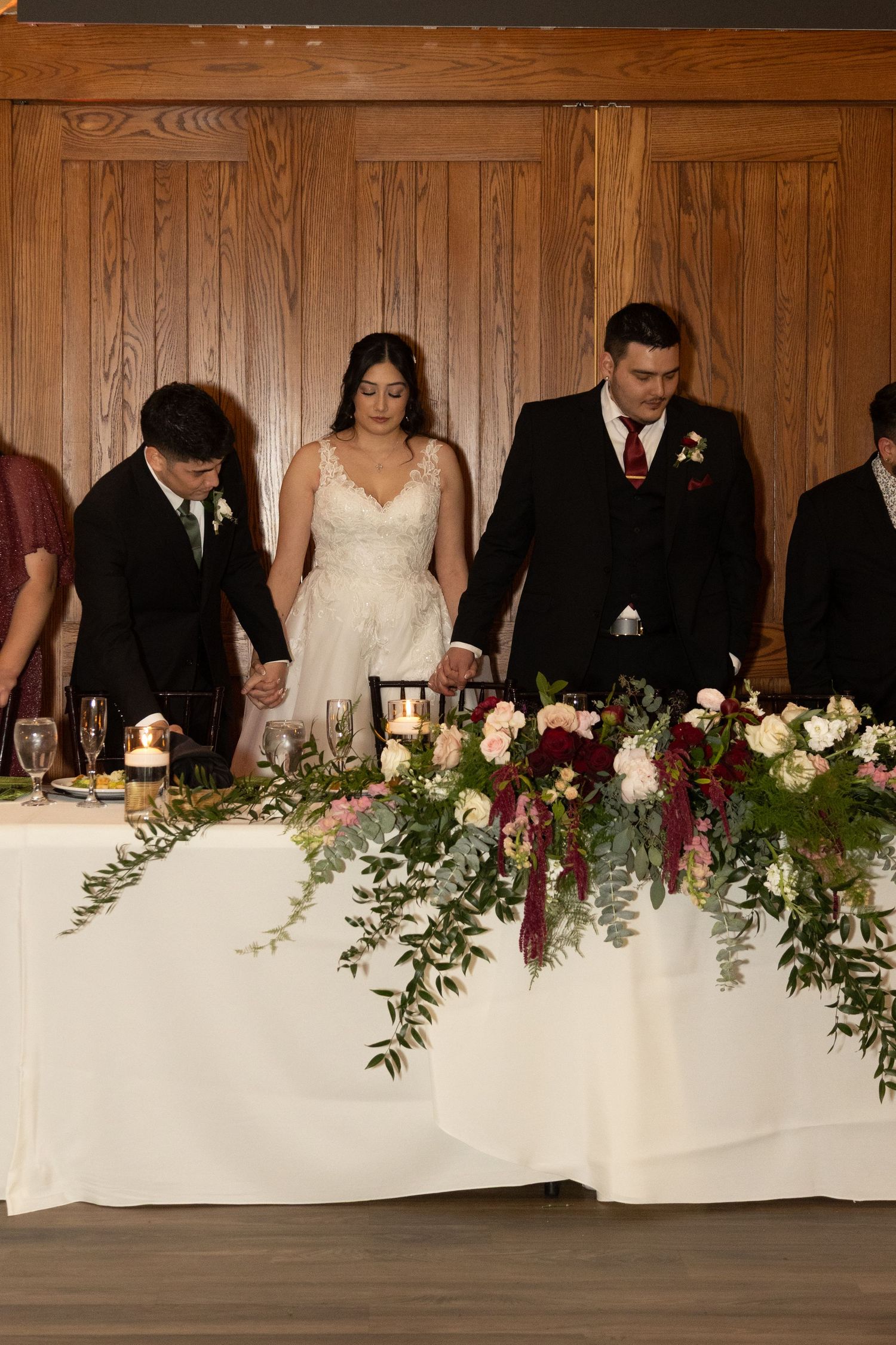 Bride and groom bow in prayer at floral-adorned head table during wedding reception.